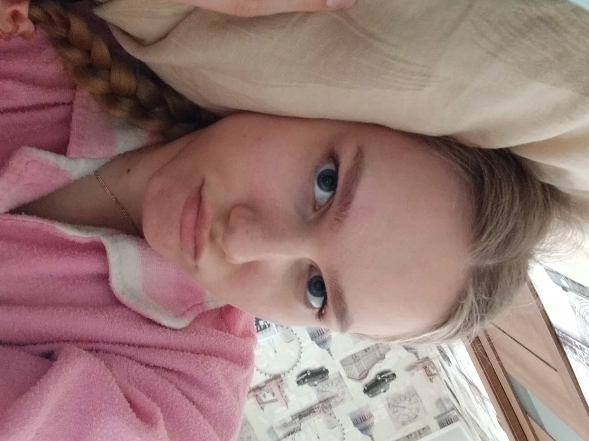 A young woman with fair skin and blue eyes is lying on what appears to be a bed or a similar soft surface. Her face is partially covered by a light-colored pillow. She is wearing a pink robe with white trim. Her blonde hair is partially braided. The surface underneath her is decorated with a pattern depicting London landmarks, including Big Ben and a taxi. The image does not provide enough information to determine if the location is Bolshaya Sosnova, Russia.