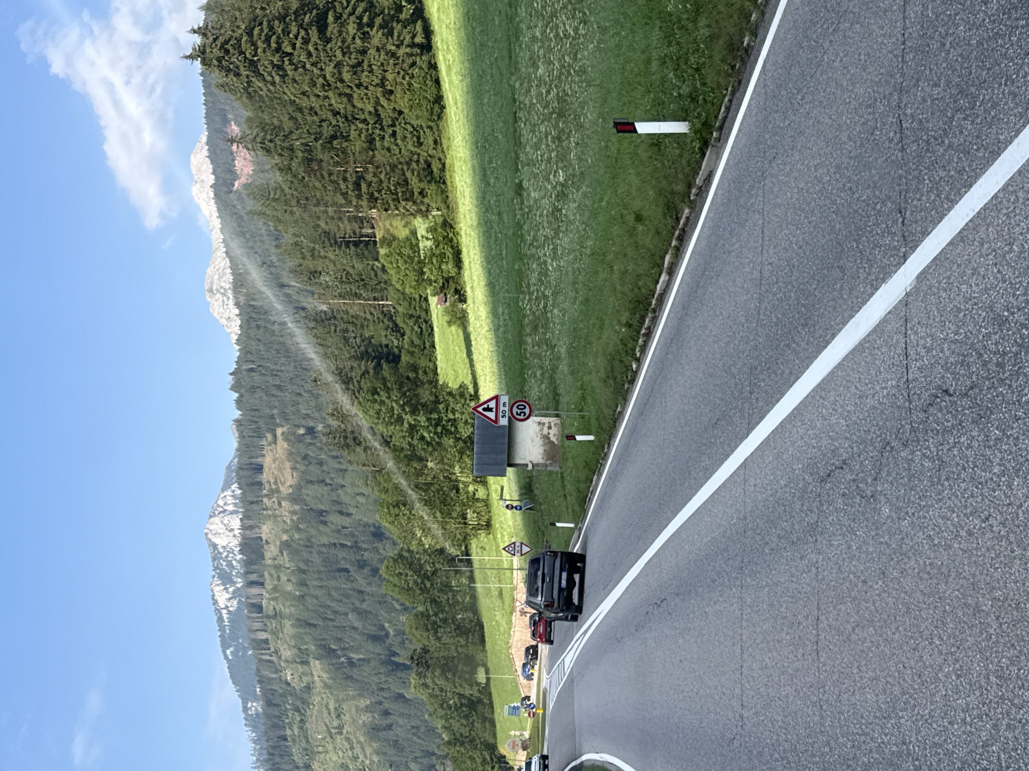 The image depicts a scenic view of a road winding through a mountainous, forested region in Vigo di Fassa - Vich, Italy. In the foreground, the asphalt road is marked with a clear white line dividing the lanes. A dark-colored SUV is traveling on the road, followed by other vehicles. The road is bordered by a grassy embankment and trees.
To the right of the road, a small, rustic building with a dark roof stands adjacent to a grassy field dotted with wildflowers. Several road signs are visible, including a triangular sign indicating a sharp turn and a circular sign with "50 m" and "50" below it, likely indicating a speed limit and distance. Red and white marker posts are placed along the side of the road.
In the background, lush green forests cover the rolling hills and steep mountainsides. The peaks of the mountains are visible in the distance, partially covered in snow, under a clear blue sky with a few wispy clouds. A subtle rainbow-like light effect is visible in the sky above the trees, possibly caused by sunlight interacting with atmospheric conditions. The overall scene conveys a sense of natural beauty and a peaceful, rural setting.