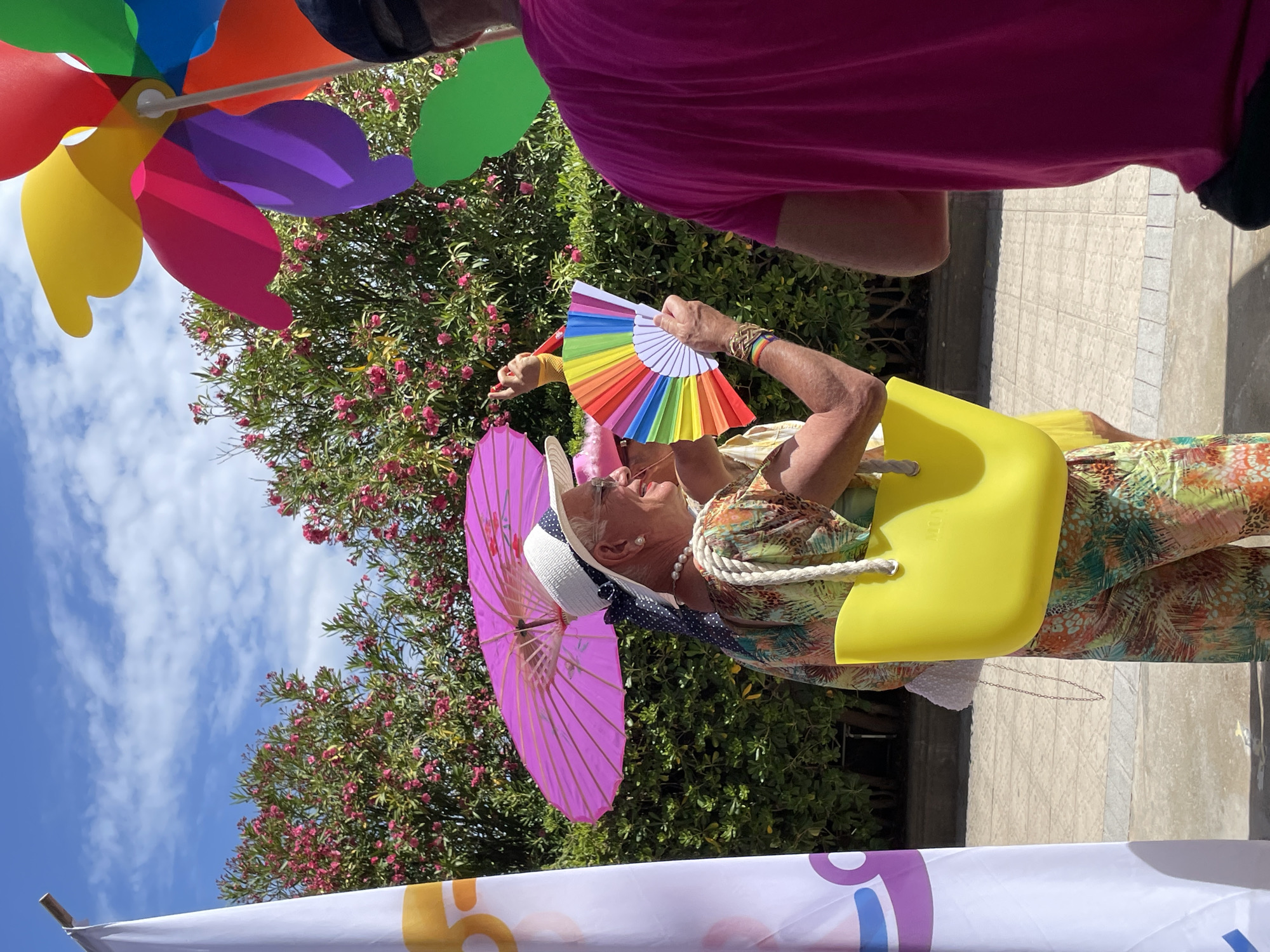 In Sitges, Spain, on a sunny day with a bright blue sky and scattered clouds, two individuals are visible outdoors. The person in the foreground, wearing glasses, a white hat with a blue polka dot ribbon, and a colorful patterned outfit, holds a vibrant rainbow fan. This individual is also carrying a bright yellow bag. Behind them, a pink umbrella is held aloft. To the right, another person is seen from behind, wearing a bright pink shirt. A colorful pinwheel is positioned in the upper right quadrant of the image. The background features lush green foliage with pink flowers. A white flag with orange and purple graphics is on the left side of the frame. The ground appears to be paved with light-colored tiles.