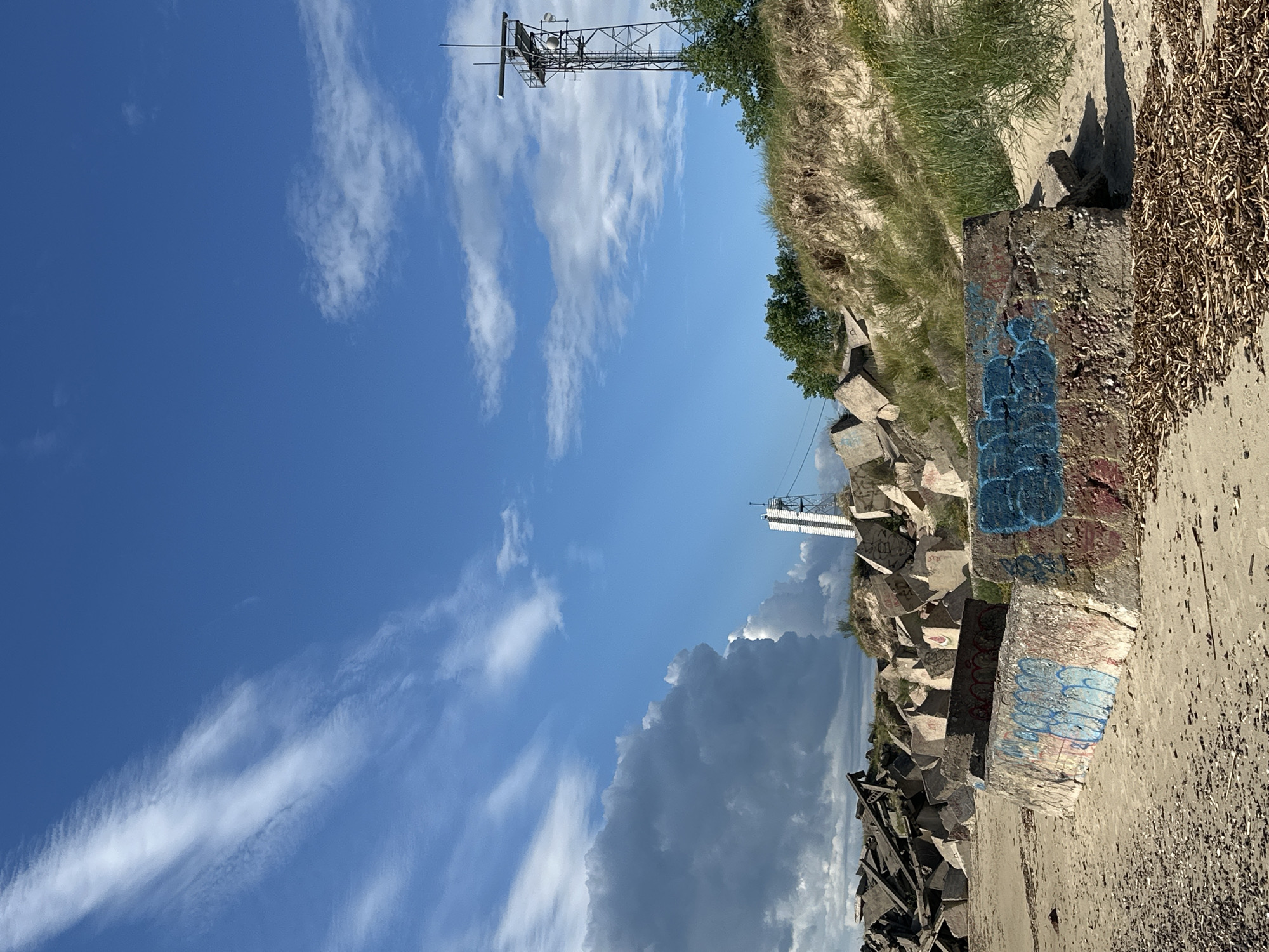 The image depicts a scene on a sandy beach, likely in Liepāja, Latvia, featuring a collection of concrete structures, some of which are covered in graffiti. In the foreground, a large, low-lying concrete block is spray-painted with blue and red graffiti. Behind this, a mound of sand rises, topped with green vegetation and a tall, dark metal tower with a radar dish at its apex, positioned to the right of the frame. Further left, a collection of angular, broken concrete blocks, reminiscent of a breakwater or defensive structure, are piled haphazardly. A tall, white, angular structure with a red top is visible in the distance, between the breakwater and the sand dune. The sky above is a clear blue, with streaks of white clouds on the left and a few scattered clouds towards the right. The ground is sandy, with scattered debris, possibly dried seaweed or wood, in the foreground. There are no people visible in the image.
