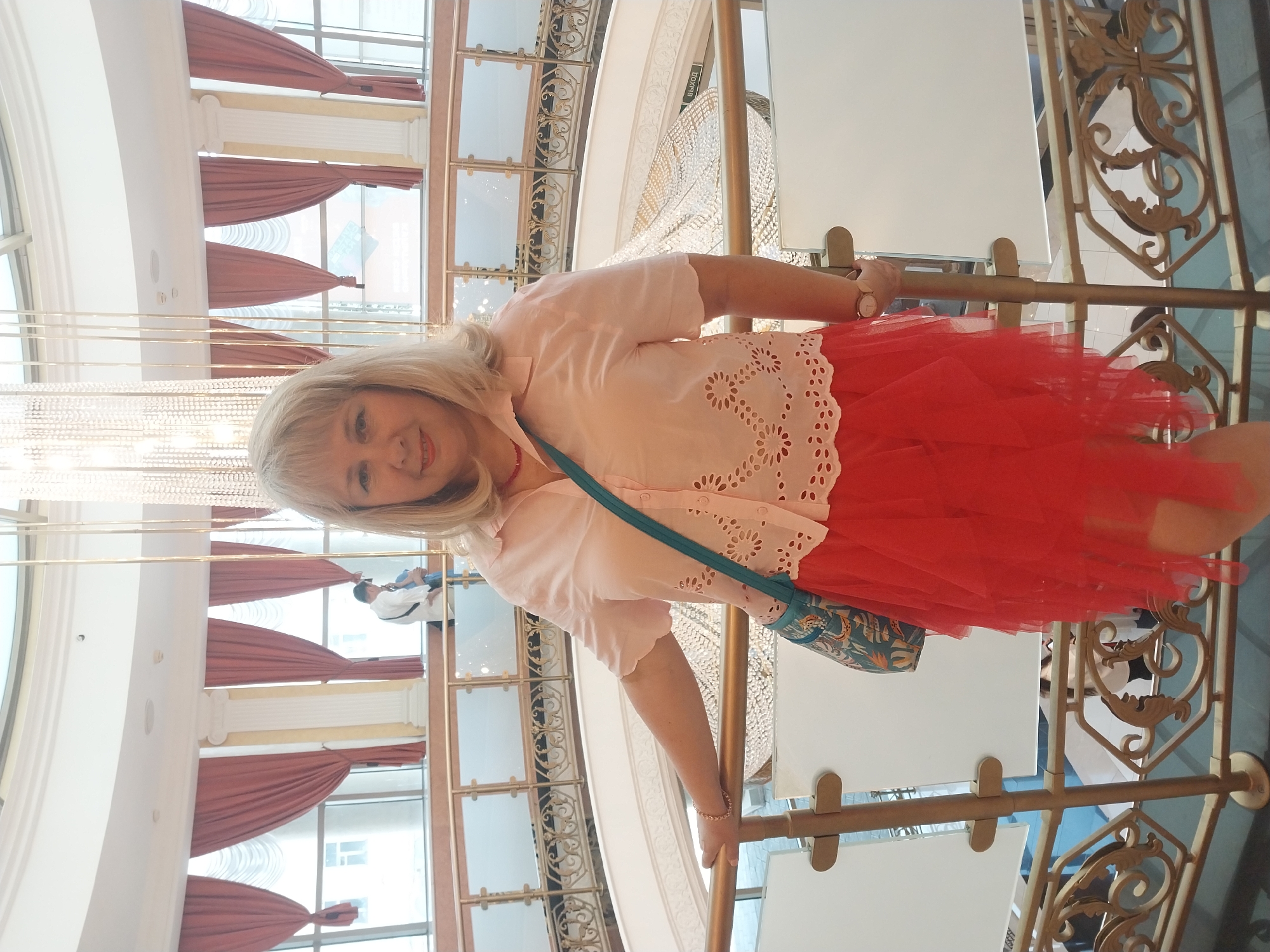 A woman with blonde hair, wearing a pink short-sleeved blouse with eyelet details and a red layered skirt, poses for a photo in an indoor atrium. She is standing next to a railing with ornate gold detailing, looking at the camera with a slight smile. A patterned teal crossbody bag is slung over her shoulder. In the background, a large, ornate chandelier hangs from the ceiling, and arched windows with red drapery are visible. Other people can be seen in the distance on a lower level. The environment appears to be a public indoor space, possibly a hotel, mall, or cultural center, suggesting it could be a location within Orenburg, Russia.