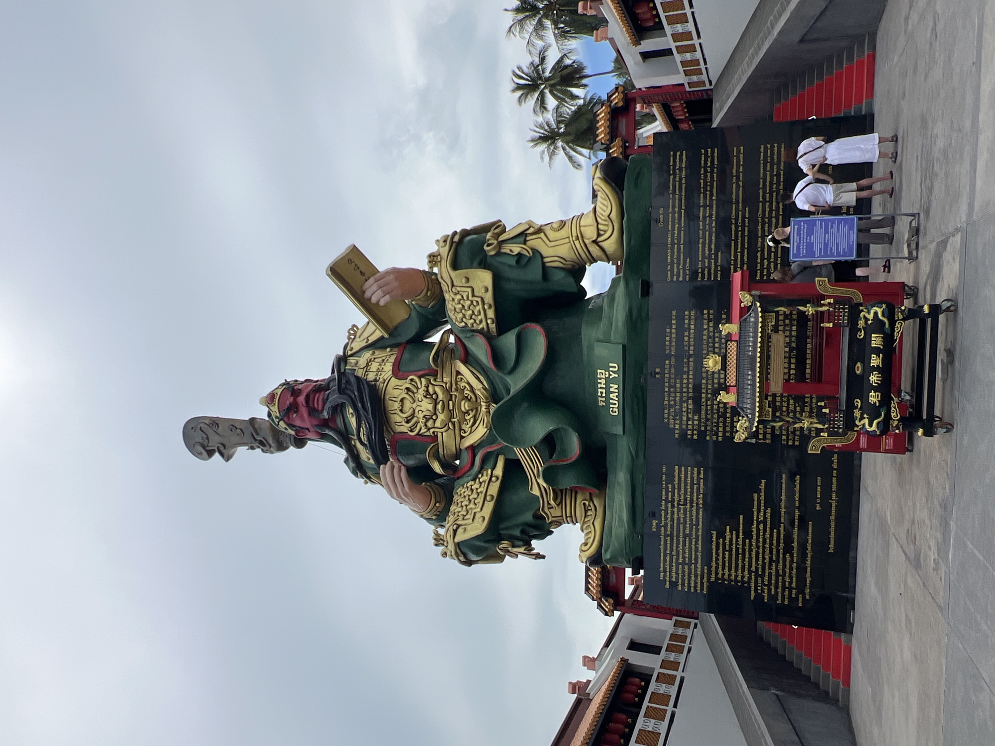 The image captures a massive statue of Guan Yu, a revered Chinese historical figure, situated in Baan Hua Thanon, Thailand. The statue is depicted in a seated posture, holding a book in its left hand and a large weapon over its right shoulder. The statue is adorned in traditional green and gold attire, with a fierce red face and a long black beard.
At the base of the statue, a large black monument displays inscriptions in multiple languages, including Thai and Chinese, detailing Guan Yu's life and significance. To the left and right of the statue, traditional Chinese-style buildings with red roofs and decorative elements are visible, flanked by red-carpeted stairs leading upwards. In front of the monument, a small, ornate altar or shrine with intricate carvings and a yellow roof is placed.
In the foreground, three individuals are standing near a blue informational sign, observing the monument. The sky above is overcast, with scattered clouds. Coconut palm trees are visible in the background, suggesting a tropical setting. The overall scene conveys a sense of cultural reverence and historical significance.