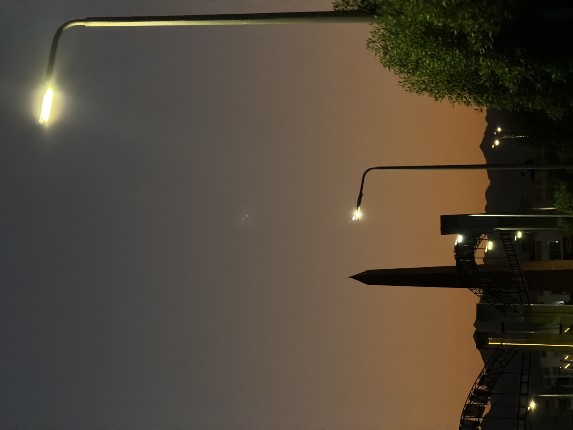 The image depicts a twilight scene in Sharm El Sheikh, Egypt. A tall, dark obelisk stands prominently in the foreground, silhouetted against the dusky orange and gray sky. To the left of the obelisk, a section of an illuminated, arched structure, possibly part of a bridge or walkway, is visible. Several streetlights are present, with one tall streetlight on the right casting a bright yellow glow, and another smaller one in the middle ground. A lush green tree occupies the far right side of the frame. In the background, mountains are faintly visible, also silhouetted against the sky. The overall impression is one of a city transitioning from day to night, with artificial lights beginning to illuminate the landscape. There are no people or animals visible in the frame.