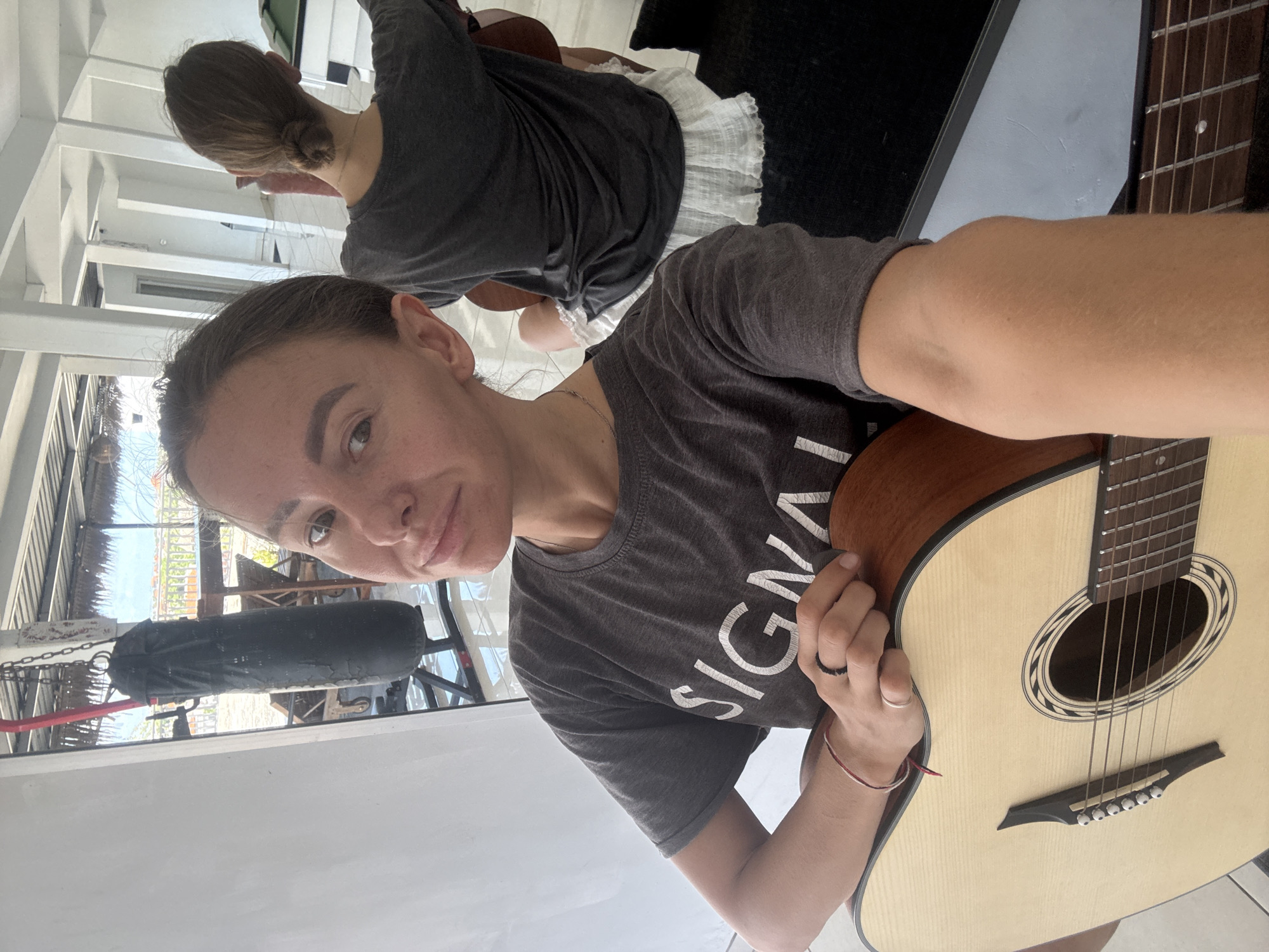 A woman with light skin, light brown hair pulled back, and wearing a charcoal grey t-shirt with white lettering is holding a light wood acoustic guitar. She has a ring on her finger and a thin bracelet on her wrist. A reflection shows her from another angle with her hair in a bun. Outside, a black punching bag hangs from a metal frame. The location appears to be in North Kuta, Indonesia.