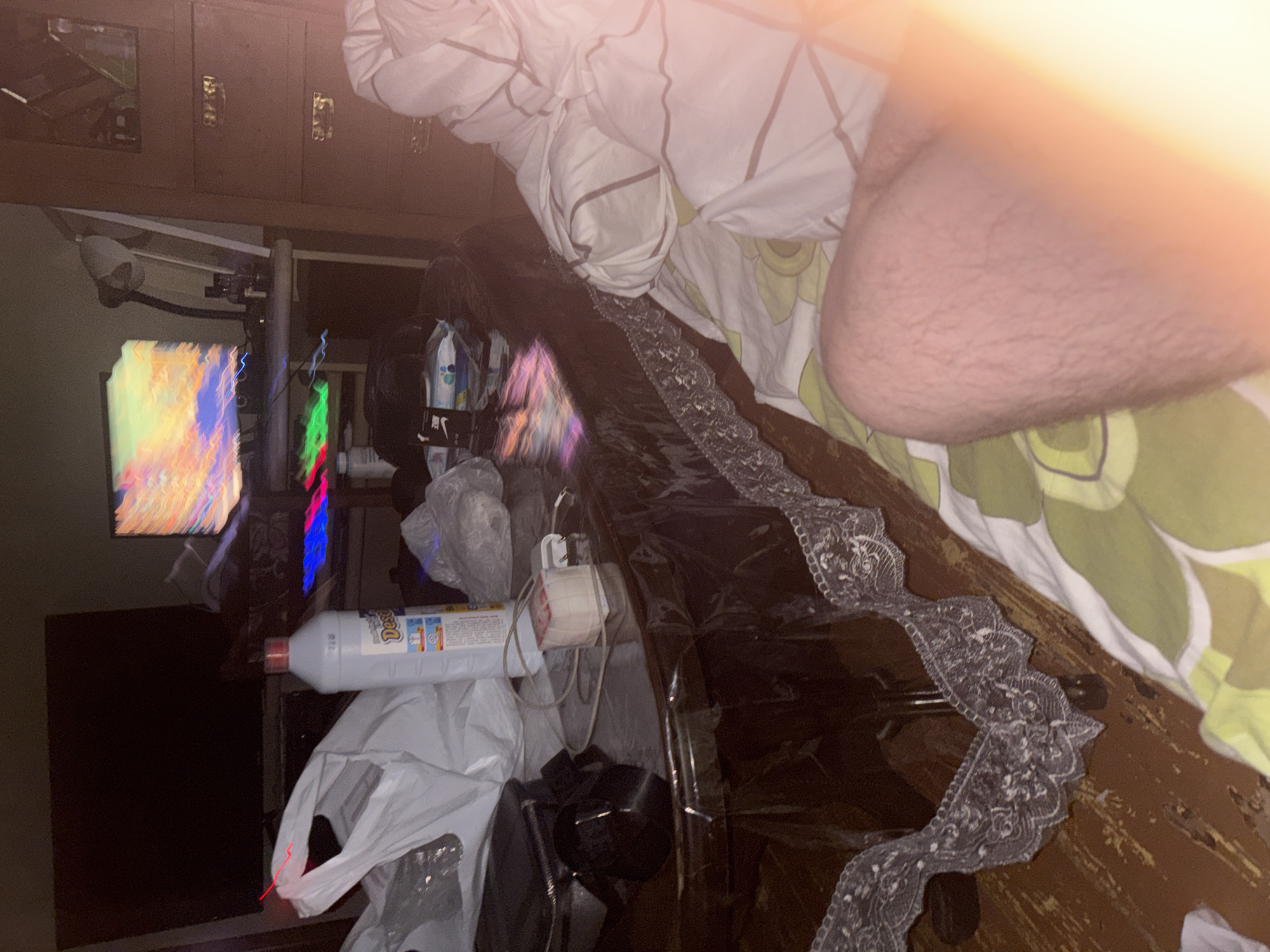 The image depicts a cluttered interior, possibly a bedroom, with a desk and bedding as prominent features. In the left side of the frame, there is a computer monitor displaying a vibrant, abstract image. To the right of the monitor, a structure with colorful LED lighting can be seen. A bottle of cleaning solution and plastic bags are scattered near the desk area. The right side of the image shows a bed with patterned bedding and a partial view of a person's arm or leg. The environment suggests a lived-in space with potential disorganization. The location in Yerevan, Armenia is not evident from the image.