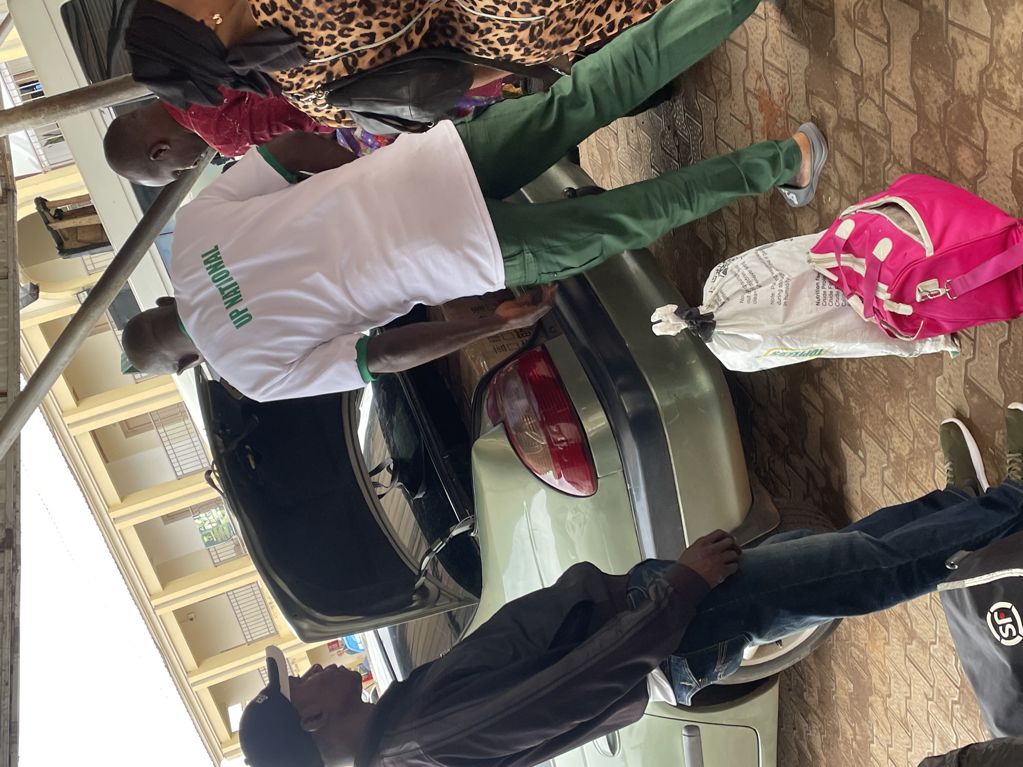 The image depicts an outdoor scene, likely in Bukuru, Nigeria, where several individuals are interacting near the open trunk of a light green sedan. The car's trunk is filled with various items, suggesting loading or unloading is in progress.
In the foreground, a man wearing a dark jacket, blue jeans, and a black baseball cap is standing and looking into the trunk. To his right, another man in a white t-shirt with green trim and the text "UP NATIONAL" printed on the back, and green trousers, is also looking towards the trunk. A third person, partially obscured, is wearing a red patterned shirt. To the right, a woman in a leopard print dress and a black headscarf is visible, carrying a black backpack.
On the ground near the car, there are two bags: a large white bag, likely containing goods, and a bright pink duffel bag. The ground surface appears to be paved with rectangular tiles. In the background, a building with yellow walls and balconies is visible, suggesting a residential or institutional setting. The overall atmosphere appears casual, with the individuals engaged in a task related to the vehicle.
