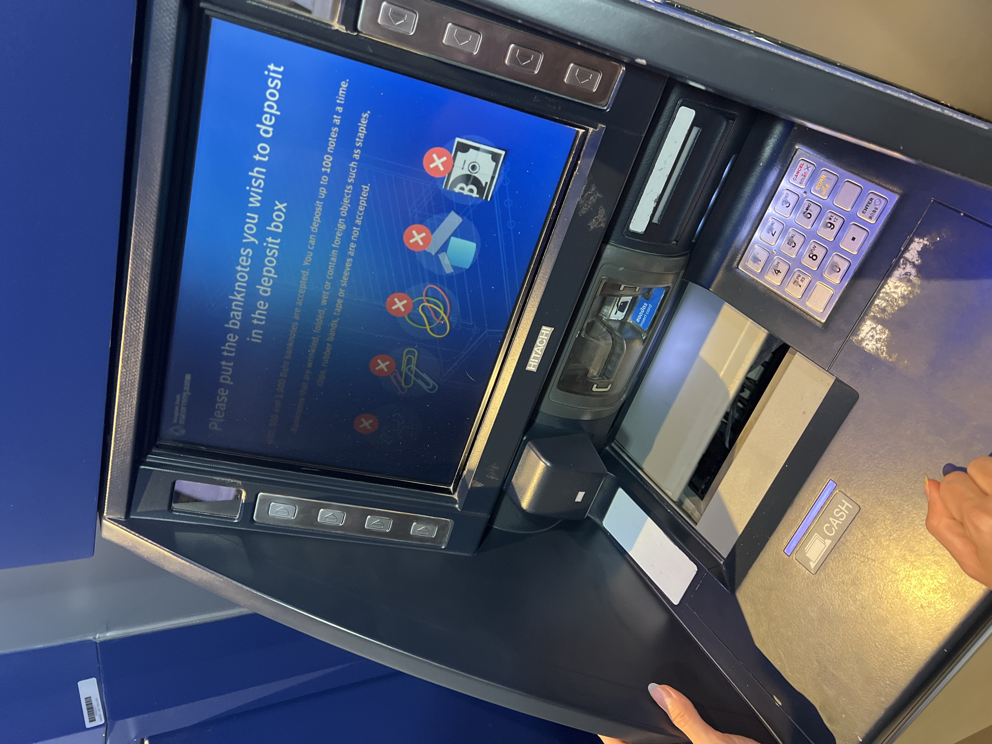 The image shows a person using an ATM in Baan Bo Phut, Thailand, to deposit cash. The ATM screen displays instructions for depositing banknotes, noting that up to 100 notes can be deposited at a time, and that wrinkled, folded, wet banknotes, or banknotes with foreign objects like staples, clips, rubber bands, tape, or sleeves are not accepted. The ATM also indicates that 500 and 1,000 Baht banknotes are accepted. The person's hands are visible near the cash deposit slot. The ATM has a keypad and a card reader. The brand of the ATM is HITACHI.