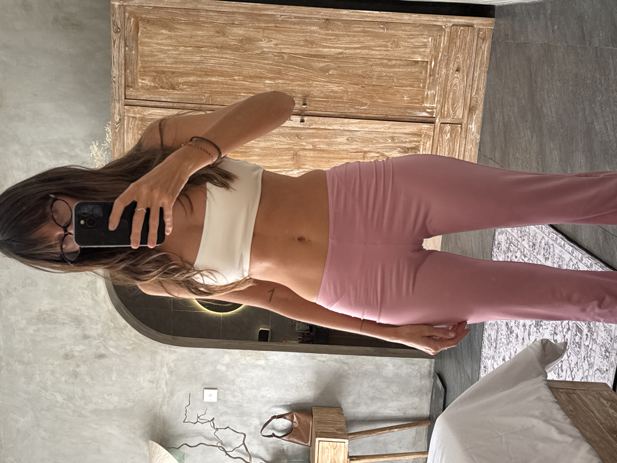 A woman with dark hair and glasses is taking a selfie in a bathroom. She is wearing a white top and pink pants. The background includes a gray wall, a wooden cabinet, a rug with a pattern, a bed, and a round mirror with a light. A purse sits on a wooden table. Based on the location tags of similar images, this may be located in North Kuta, Indonesia.