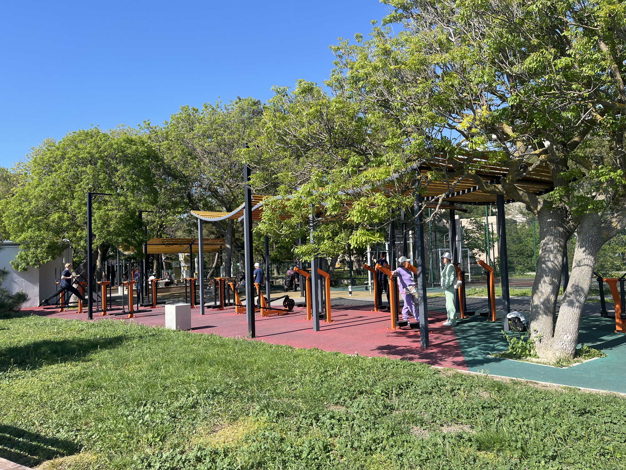The image shows an outdoor exercise area in Anapa, Russia, during daylight. Several people, possibly elderly, are using the exercise equipment. The area has a red and green rubber surface, with exercise machines consisting of black metal frames and orange supports. A pergola structure with wooden slats provides partial shade. Trees with green foliage surround the exercise area. In the foreground, there is a patch of grass. The sky is clear and blue.