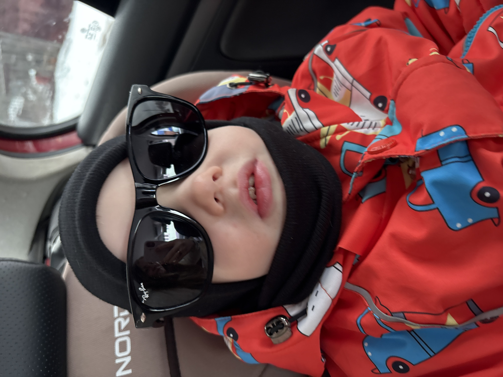 A child wearing a black beanie and oversized Ray-Ban sunglasses is lying in a car seat. The child is wearing a red jacket with a pattern of cartoon cars. The car seat is beige and black, and part of the word "NORDI" is visible on the headrest. The car's interior is partially visible, including the window. The location is Chaikovsky, Russia.