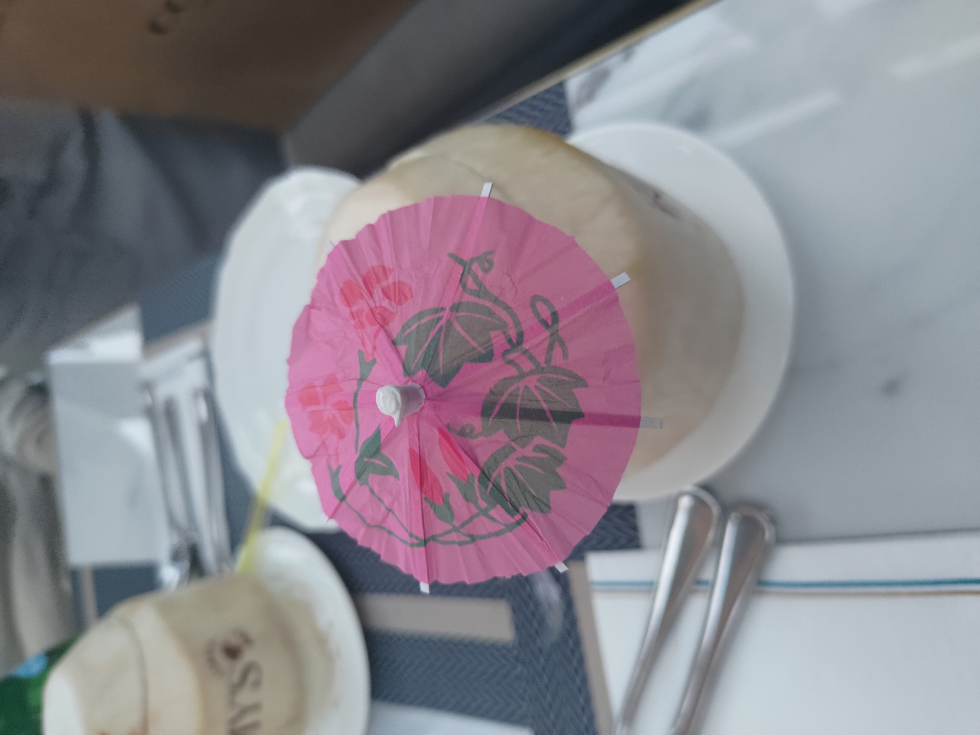 In the city of Kosambi Barat, Indonesia, a close-up, high-angle shot captures a table setting for a meal, possibly in a restaurant. Two whole coconuts are featured, one partially visible on the left and a larger one in the center, topped with a small, pink paper umbrella decorated with floral patterns. The coconuts sit on white plates, which are placed on a dark blue placemat with a striped pattern. In the background, cutlery, including two forks, is laid out on a white surface. The overall environment appears to be indoors, with soft lighting suggesting a casual dining atmosphere.