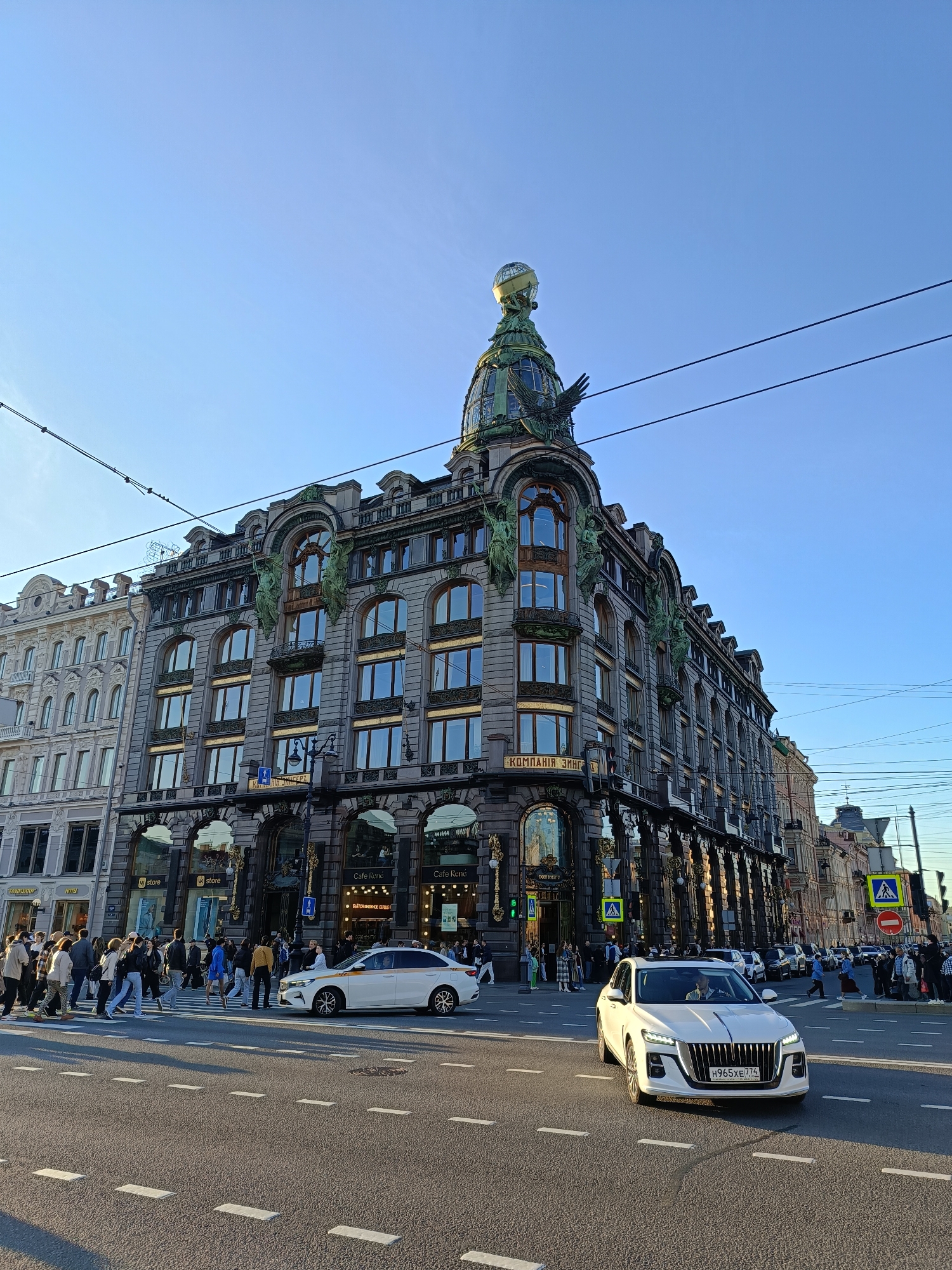 The image shows the corner of the Eliseyev Emporium building in Saint Petersburg, Russia, taken from a street-level perspective. The building, known for its ornate architecture, occupies the corner of a busy intersection. People are crossing the street at a crosswalk in front of the building. Two white cars are in the foreground, one of which has license plate “H965XE 7.74”. The building has multiple stories with arched windows and is adorned with statues. The dome of the building is visible at the top, featuring a sculptural globe. The sky is clear and blue. Utility wires are strung across the upper part of the frame. "KOМПАНIЯ ЗИНГ" and "БЪЕТСЯ КНИЖНОЕ СЕРДЦЕ" are partially legible on a sign affixed to the building's exterior. Storefront signs reading "store" and "Cafe Rene" are also visible.
