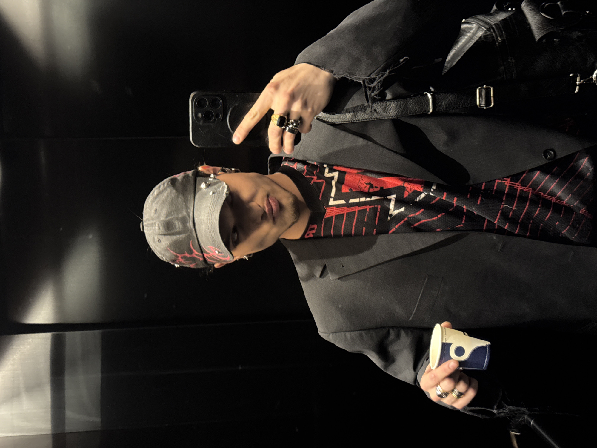 A young adult male is captured in a selfie within a reflective, dark-paneled interior, identified as likely an elevator in Sofia, Bulgaria, based on the branding on a disposable cup. The subject faces the camera directly, holding a black smartphone vertically in his right hand to take the photograph. His left hand holds a small, blue and white paper cup with a stylized graphic resembling a 'G' or coffee bean, consistent with Geshkov Coffee Roasters, a business located in Sofia.
The male wears a distressed grey baseball cap featuring red flame-like designs and several silver studs/spikes on the left side. He has a short goatee and a hoop earring in his visible left ear. His attire includes a dark, possibly black, blazer with noticeable fraying and damage on the cuffs and right shoulder, worn over a long-sleeved shirt with a black base and red and white geometric patterns. Multiple rings adorn his fingers: a large black and gold ring on his right ring finger, and two stacked black and silver rings on his right middle finger. A black leather-like shoulder bag with buckles and distressed elements is worn over his left shoulder. The background exhibits subtle reflections and vertical lines consistent with elevator panel seams.