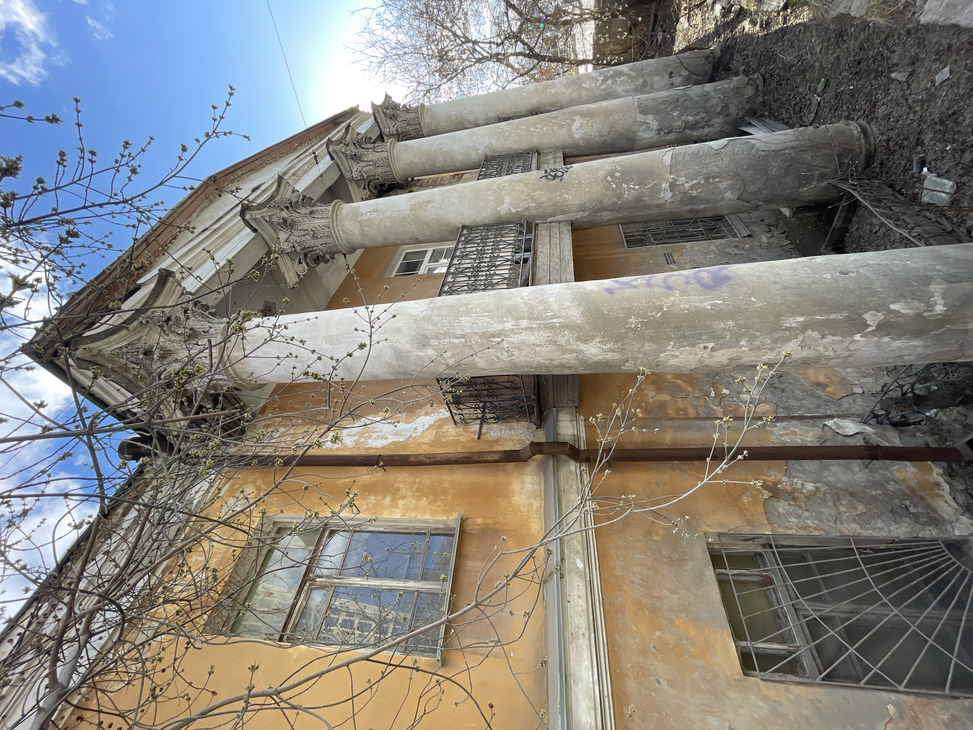 An exterior shot of the abandoned Sevastianov Estate in Yekaterinburg, Russia, reveals a deteriorated, three-story building. The facade is a mix of peeling, yellow-toned stucco and crumbling, gray-white columns. Ornamental ironwork adorns the windows, and the columns are capped with weathered, decorative elements. A bare tree with budding branches extends across the lower left portion of the frame. The sky above is a bright blue, with a hint of glare. The building is tilted at an angle, as if viewed from below. The top of the building is cluttered with debris, and the structure shows signs of neglect.