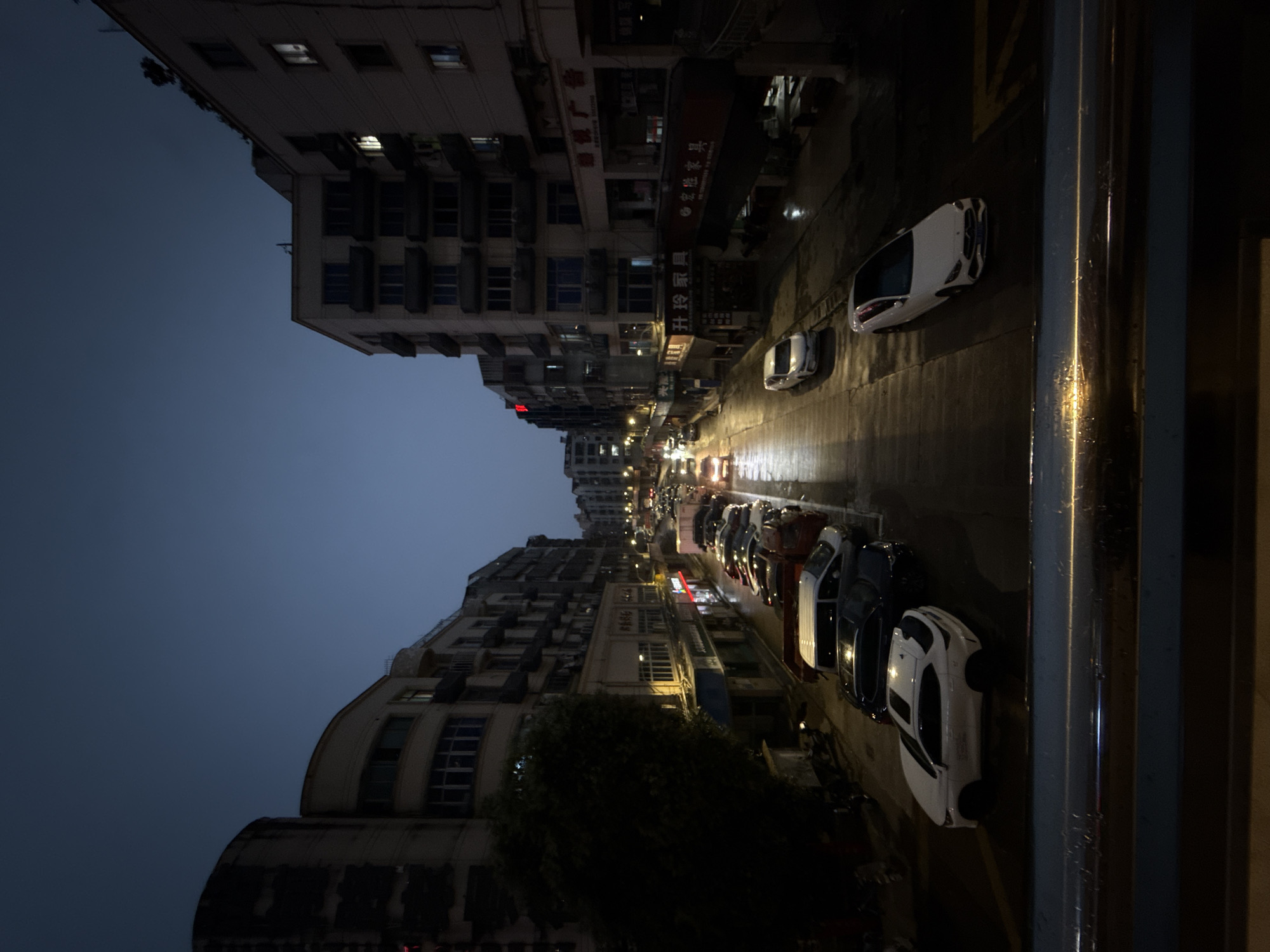 The image depicts a street scene in Yiwu, China, during twilight. The sky is a deep blue, transitioning from the fading daylight. The street is lined with multi-story buildings on both sides, characterized by balconies and numerous windows, some of which are illuminated. The architecture suggests a mix of residential and commercial spaces.
A considerable number of cars are parked along the sides of the street and some are moving in traffic. The street appears to be wet, reflecting the streetlights, indicating recent rain or a damp surface. The lighting in the scene is dim, primarily from streetlights and the lights within the buildings, creating a somewhat moody atmosphere.
On the left side, a large, dark tree stands near the buildings. On the right side, store signs are visible, some featuring Chinese characters, suggesting a commercial area. The overall impression is of a typical urban street in China as evening sets in.