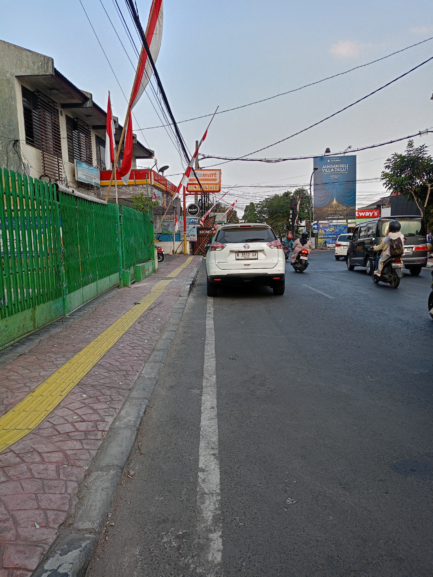The image depicts a street scene in Batu, Indonesia, likely on a sunny day with a clear sky. A white SUV, license plate "N 1512 LF", is parked on the right side of the road, facing away from the viewer. In front of the SUV, a dark-colored van with license plate "JU" is also visible, as are several motorcycles, some carrying passengers. Red and white Indonesian flags are prominently displayed, some attached to poles extending over the street, and others on buildings.
To the left of the road is a sidewalk paved with textured red bricks, bordered by a yellow tactile paving strip. A green metal fence runs along the edge of the sidewalk, adjacent to a building with shaded windows. Signs for "Alfamart" and a billboard for "Toweland JANGAN BELI VILLA DULU!" are visible in the background. The road itself is asphalt, with white lane markings. The overall impression is a busy urban street with typical traffic and commercial elements.