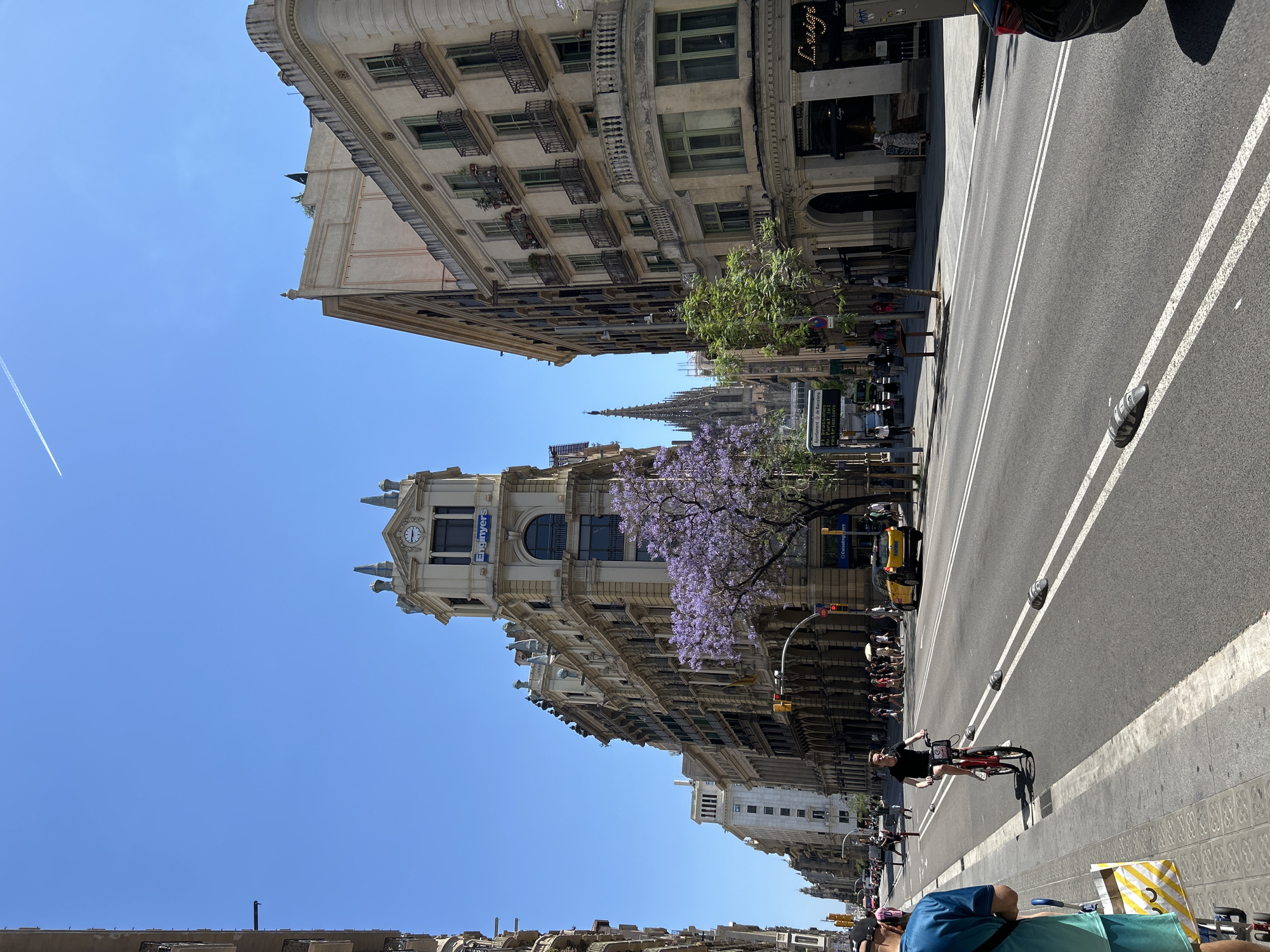 The image captures a street scene in Barcelona, Spain. The street is lined with grand, ornate buildings, including the Col·legi d'Enginyers Industrials de Catalunya building, identifiable by its clock tower and signage. A mature Jacaranda tree with purple blossoms adds a vibrant splash of color. Pedestrians and cyclists are present, with one cyclist in the foreground on a red bicycle. Cars and taxis can be seen in the street. The sky is a clear blue with a contrail visible. The general environment suggests a busy, urban thoroughfare.