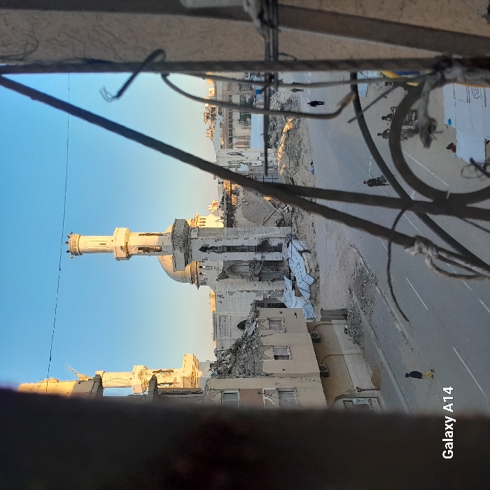 The image displays a scene of significant urban destruction centered on the Great Mosque of Khan Yunis, Palestinian Territory. The photograph is taken from an elevated position, framed through a window or opening with dark, irregular metal bars and wires partially obscuring the lower and right foreground.
In the background, the Great Mosque of Khan Yunis stands with its central dome largely intact but its main facade severely damaged, showing large sections of missing walls and exposed structural elements. Rubble is piled directly in front of the mosque's entrance. Two tall, white minarets with circular upper sections flank the mosque; the left minaret shows some damage, while the right one appears relatively intact from this angle.
To the left of the mosque, a multi-story building exhibits extensive damage to its upper floors, with exposed rebar and concrete debris. White tarpaulins or sheets are visible covering some of the rubble piles in front of this building. To the right of the mosque, further structural damage and debris are visible, extending into the distance where less damaged buildings can be seen under a clear blue sky.
In the midground, a paved road with a solid white line and a dashed white line runs horizontally. The road appears clear of major debris but is flanked by significant amounts of rubble. Several individuals are present on and near the road:
* One individual in a yellow shirt and dark pants walks left on the paved road in the lower-left quadrant.
* Two individuals in dark clothing walk away from the viewer along the right side of the road in the mid-distance.
* On the far right, near the partially obscured foreground, a small group of 2-3 individuals are gathered near piles of white-covered materials or debris, one appearing to push or interact with a small cart or trolley.
The scene is illuminated by natural light, casting shadows from the left, highlighting the front of the mosque and minarets. The overall environment indicates widespread devastation consistent with conflict or severe structural damage. "Galaxy A14" is watermarked in the lower-left corner.