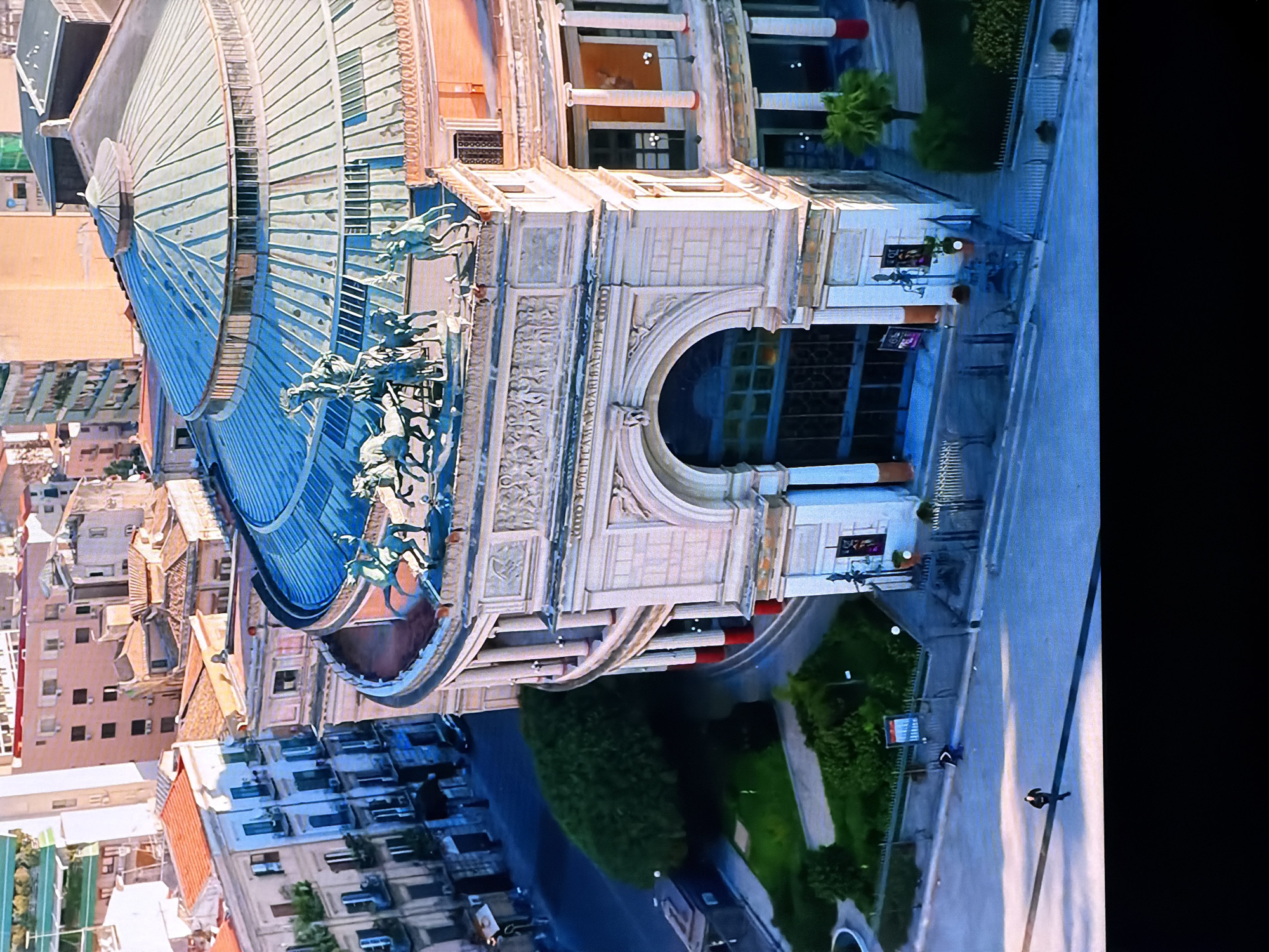 The aerial image shows the Teatro Massimo in Palermo, Italy. The opera house is a grand neoclassical building with a large, circular, domed roof made of greenish metal. Sculptures of horses and chariots adorn the top of the building. Below the dome, there is an elaborate facade with columns, arched entrances, and intricate carvings. The surrounding cityscape features a mix of residential and commercial buildings, with terracotta roofs and balconies visible. In the foreground, a street is visible with a few people walking. The image captures the historical and architectural significance of the location. The city of Chirchik, Uzbekistan is not depicted in this image.