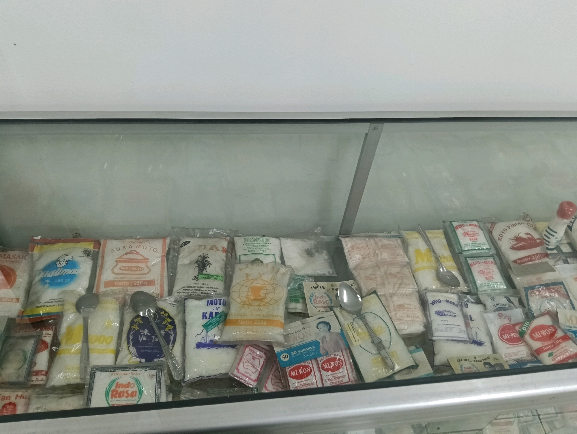 The image shows a refrigerated display case filled with various food products, primarily seasoning packets and powders. The items are mostly packaged in small, colorful bags and sachets. Some of the visible branding includes "Suka Moto," "Masak," "Indo Rasa," "Mi-WON," and "Dap-Rasa." There are also several spoons placed within the display, likely for customers to handle or for sampling. The display is well-lit, and the products appear to be neatly arranged. The background consists of a white wall and the reflective surface of the display case. The scene is set in a retail environment, possibly a small grocery store or a market stall in Kletek, Indonesia.
