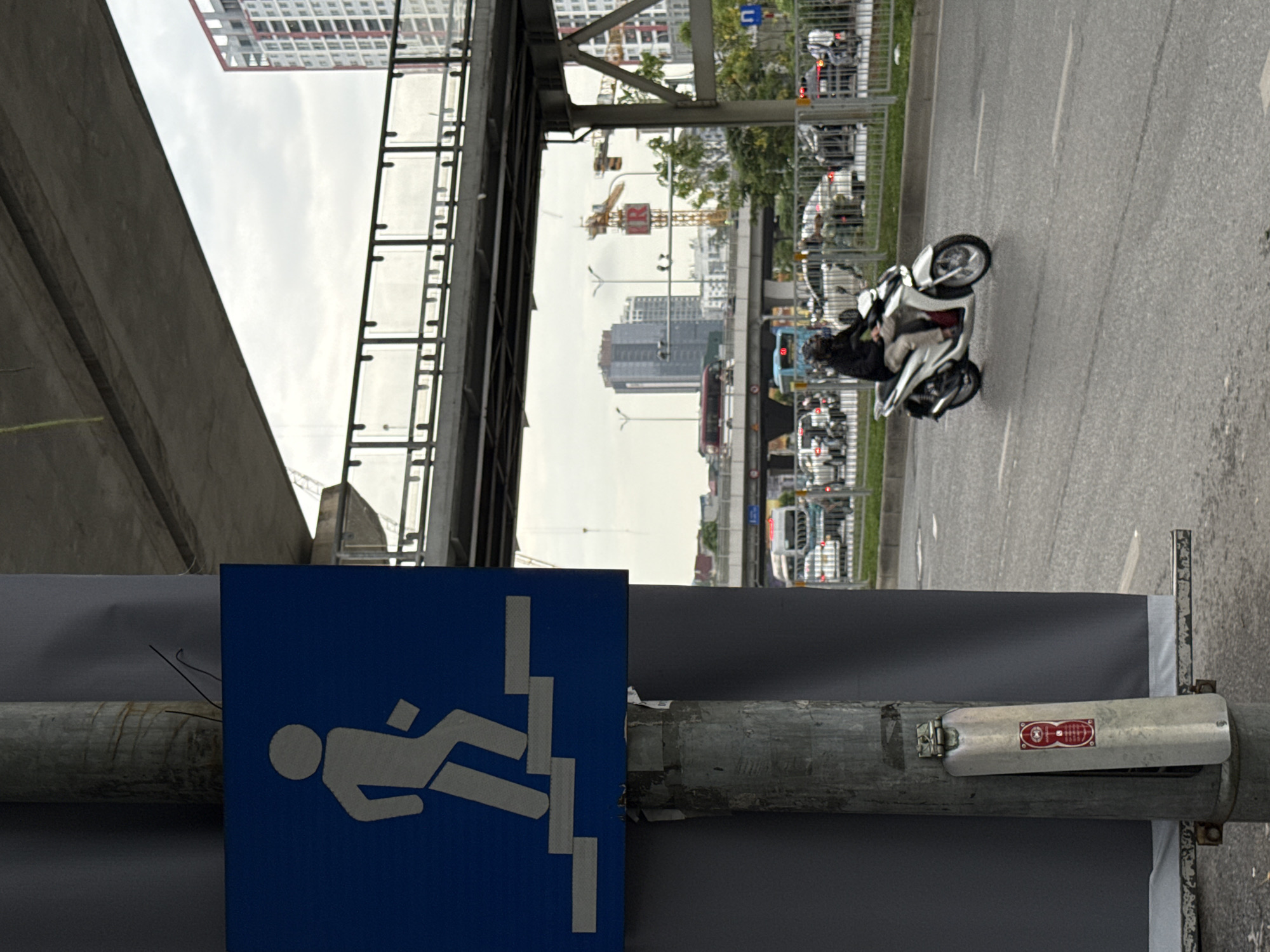 A view of a city in Hà Nội, Vietnam, featuring a pedestrian walkway sign. The sign, affixed to a weathered metal pole, depicts a figure walking down stairs. In the background, there is traffic on a road, including a person riding a white motorcycle. Overpasses, construction cranes, and buildings are also visible in the background.