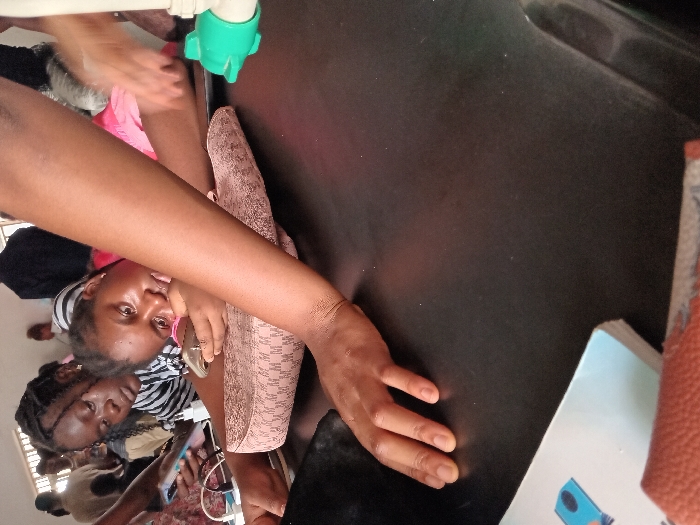 The scene is set indoors in OGBOKO, Nigeria, at what appears to be a communal or educational facility, observed from a low vantage point across a dark, smooth tabletop. In the midground, two young women are positioned. The woman closer to the right, wearing a pink top, rests her left forearm on a beige patterned purse or bag. She holds a smartphone horizontally in her right hand, her attention directed towards the device or a point just beyond it. Behind her and to the left, another woman in a black and white striped top holds a smartphone in her left hand, also looking downwards. Her hair is styled in dark braids. Multiple white charging cables and power adapters are visible on the table near these individuals, indicating device charging. A light-skinned forearm and open hand extend from the right foreground, resting on the tabletop. On the far right, a white pipe with a green connector is visible just above the table surface. In the bottom-left corner, a light blue and white book is partially visible, resting on an orange-brown textured object. The background features several out-of-focus individuals and a window with vertical bars, suggesting an interior space with ambient lighting.