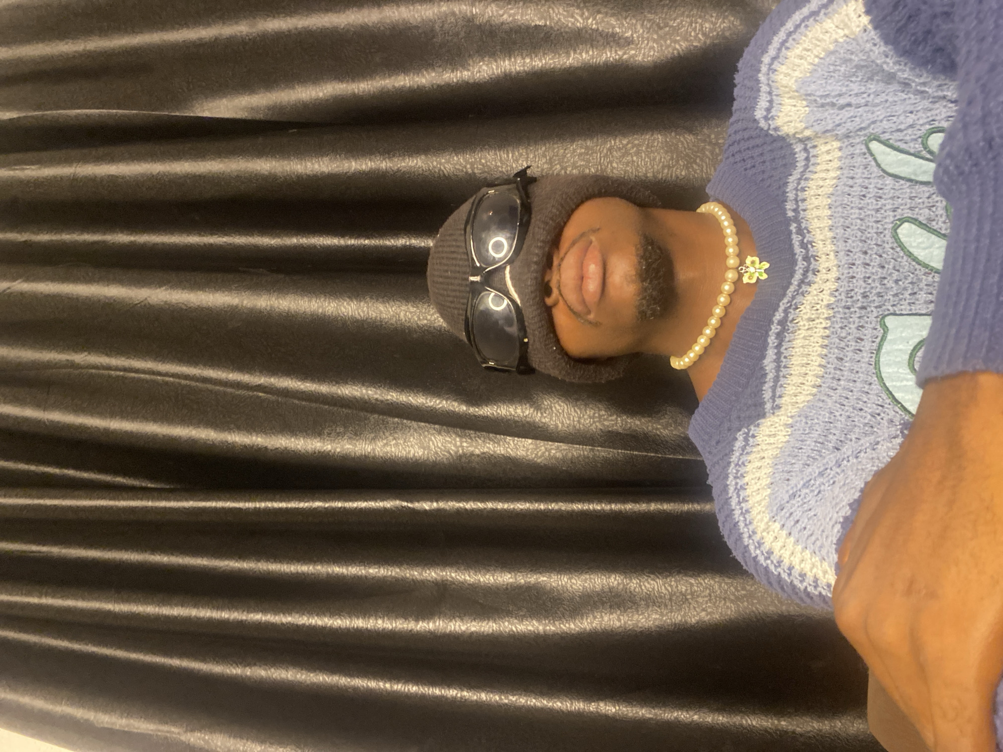 A person wearing a beanie and goggles is seen in front of a dark, textured curtain. They are also wearing a pearl necklace with a small green and yellow charm, and a blue and white striped sweater. The person's face is partially obscured by the beanie and goggles, but their lips and chin are visible. There is a faint goatee. The background appears to be a domestic setting, possibly an indoor space in Abakaliki, Nigeria, given the context provided. The lighting is dramatic, casting shadows and highlighting the textures of the curtain and clothing.