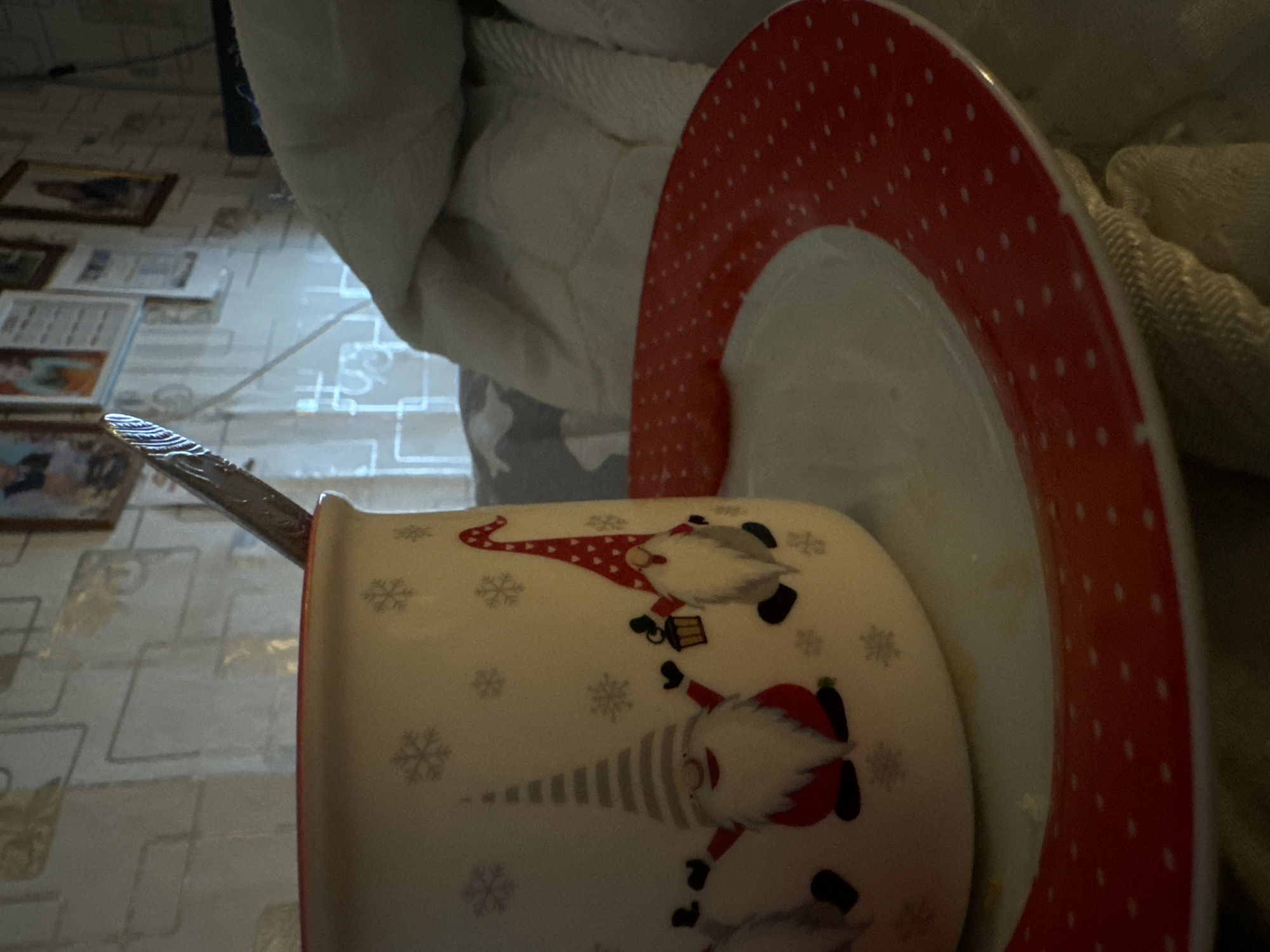 The image depicts a mug with a spoon resting inside and a plate with a red and white dotted rim placed next to it. The mug is decorated with images of gnomes and snowflakes. The scene includes a patterned surface, possibly a tablecloth, in the background. The location is unspecified.