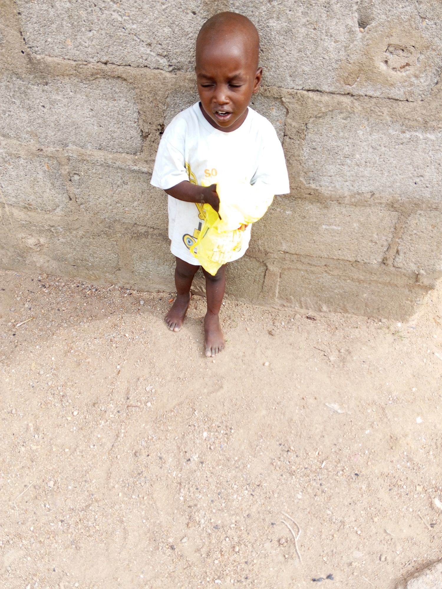 A full-shot outdoor photograph from Adonkolo, Nigeria, captures a young child, likely a boy, standing barefoot against a weathered concrete block wall. The child has dark skin and a shaved head, and wears a simple white short-sleeved t-shirt. On the upper left chest of the t-shirt, the yellow letters "SO" are visible. He is holding a crumpled yellow object, possibly a plastic bag or fabric, with both hands in front of him.
The child's facial expression suggests distress or discomfort; his eyes are partially closed and cast downwards, and his mouth is open as if he is about to speak, cry, or is experiencing an unpleasant sensation.
The background consists of the rough, light-grey concrete block wall, which appears to be constructed from large, rectangular blocks. The ground beneath the child's feet is a light sandy or dirt surface, interspersed with small pebbles and scattered debris. The lighting indicates it is daytime, likely sunny, as suggested by the bright illumination and subtle shadows. The overall scene depicts a moment of vulnerability for the child in a simple, outdoor setting.