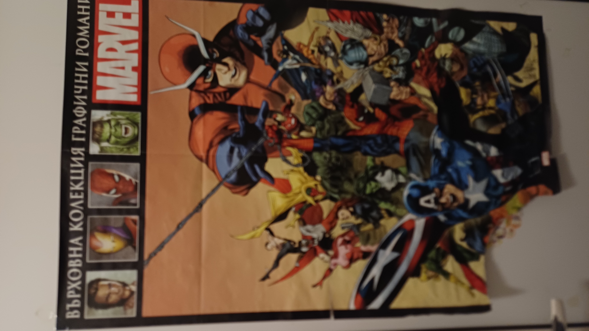 A poster is displayed on a white wall. The poster features a collection of Marvel comic book characters. At the top of the poster, text in a foreign language is visible, followed by images of Wolverine, Iron Man, Spider-Man, and the Hulk. Below these, the "MARVEL" logo is prominent. The main body of the poster depicts a large gathering of superheroes in action poses, including Captain America in the foreground on the left. The characters are drawn in a comic book style with vibrant colors against an orange background. The poster appears to be made of paper and is attached to the wall with what looks like tape. The environment is a simple interior space with a white wall.