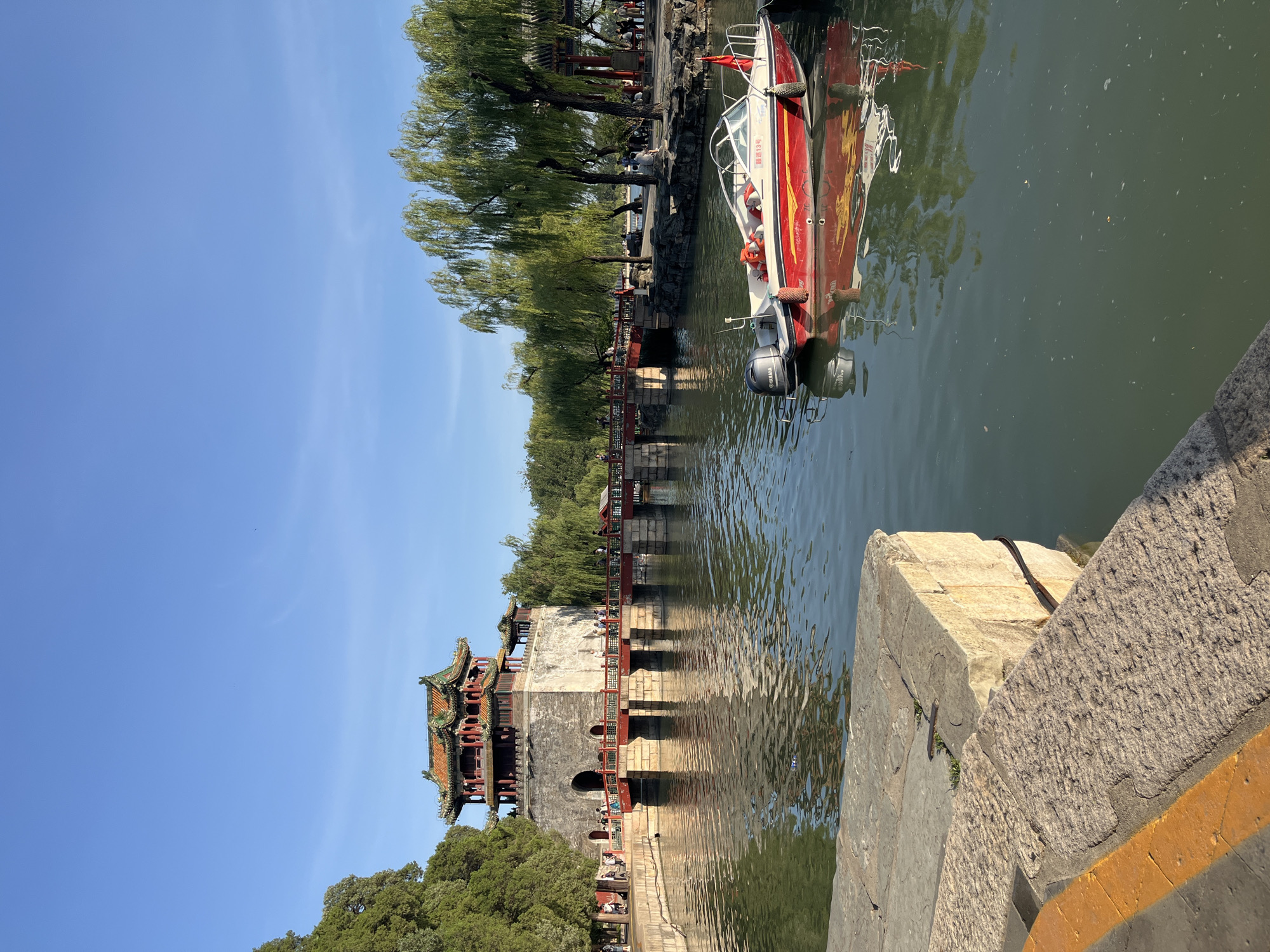 A scenic view captures a tranquil scene by a body of water, likely a lake or river. In the foreground, a stone bridge or embankment made of rough-hewn blocks leads the eye towards a traditional Chinese-style building with an ornate, multi-tiered roof featuring green tiles and red accents. Lush green trees with drooping branches, characteristic of willows, line the water's edge, casting gentle reflections on the water's surface.
A red and white motorboat is docked or slowly moving on the water, its reflection clearly visible. The boat appears to be a passenger vessel, with several people visible on its deck. The boat has an outboard motor attached to its stern. There is text on the boat, which appears to be "13号" within a yellow circle.
Several people are scattered along the waterfront and on the bridge, enjoying the scenery. The sky above is a clear, bright blue, suggesting it is a sunny day. The overall atmosphere is peaceful and serene, evoking a sense of calm and leisure. The time of day appears to be late morning or early afternoon, judging by the bright sunlight and clear sky. The architectural style of the building and the type of trees suggest a location in East Asia, possibly a park or historical site.