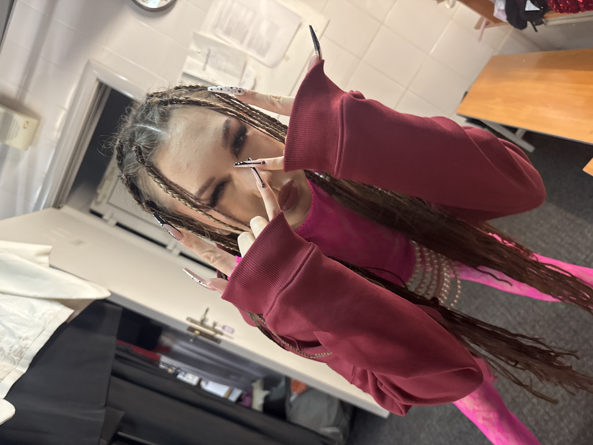 A woman with braided hair is posing indoors. She is wearing a pink top and a maroon hoodie, and pink leggings. She is making a hand gesture with long painted nails. The location is in a room with white tiled walls, fluorescent lighting, and a table and other furniture in the background. There is no evidence indicating the city of Kazan, Russia.