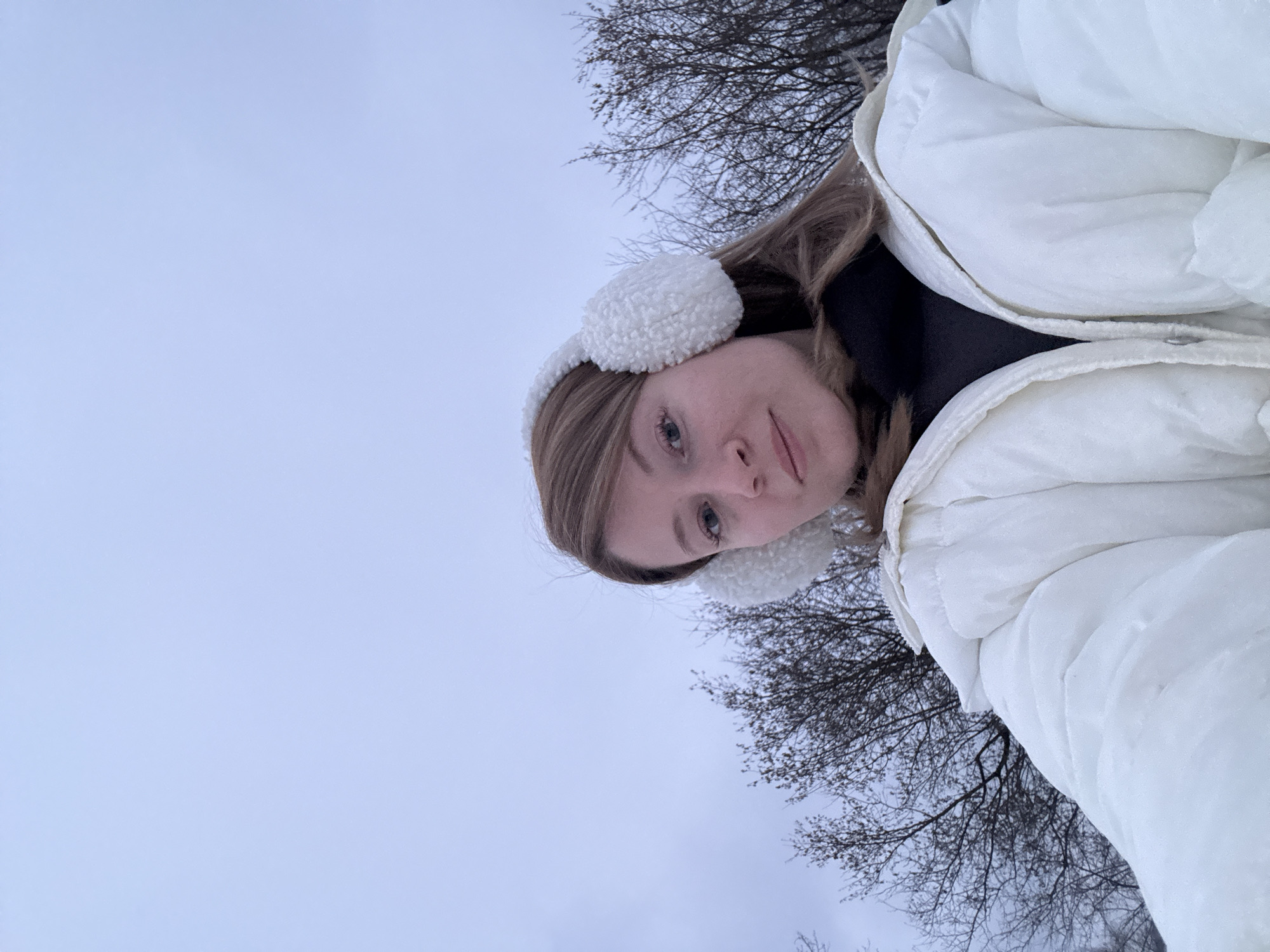 A woman wearing a white, quilted jacket and white earmuffs is outdoors. She is looking at the camera with a slight smile. Behind her are bare tree branches silhouetted against a light gray sky. The location is Вассята, Russia.