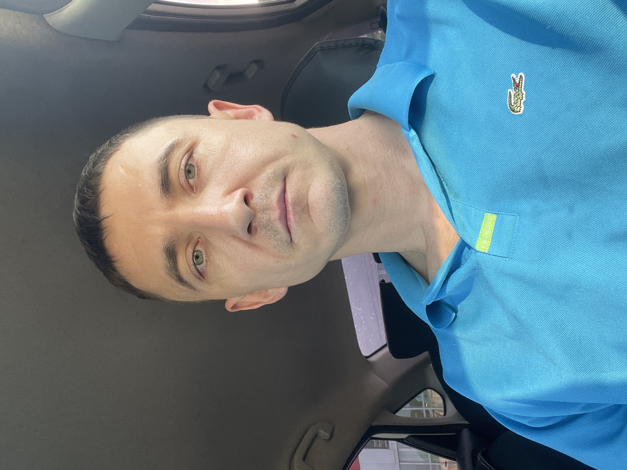 A man with short dark hair and light green eyes is photographed from the chest up, sitting in the passenger seat of a car. He is wearing a bright blue polo shirt with a green and yellow tag on the collar and a Lacoste logo on the chest. The background shows the interior of the car, including the headrest and part of the car door. Outside the car window, a building with windows is visible, suggesting an urban environment. The man's expression is neutral. The image appears to have been taken in Laishevo, Russia.