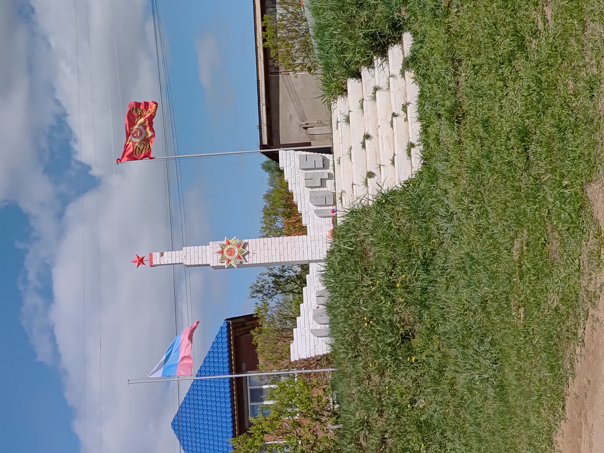 The image depicts a monument in Markovo, Russia. It features a white brick structure with the inscription "1941-1945." Above the inscription, there is a five-pointed red star and an emblem resembling a war medal. Two flags flank the monument. The upper one is red with a gold emblem, and the lower one consists of white, blue, and pink stripes. Green foliage surrounds the monument on the ground. The background includes a blue sky with white clouds, a building with a blue roof, and power lines.