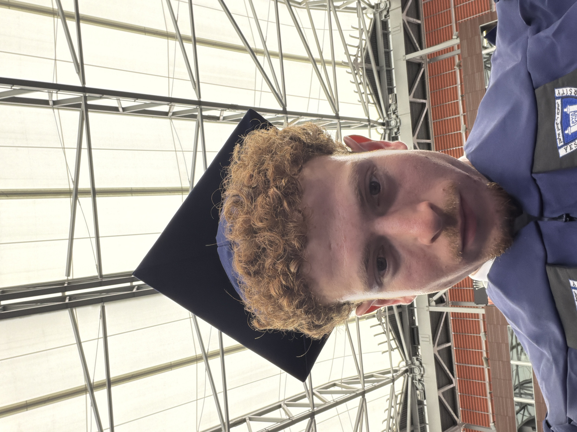 A person wearing a graduation cap and gown is standing inside what appears to be a large, modern structure with an intricate ceiling of metal beams and glass panels. The person is facing the camera. The building has windows, and there is an academic logo on the gown, with the words "Yeshiva University" visible. Although the image does not definitively confirm it, the architectural style and the presence of Yeshiva University strongly suggest that the location could be within New York City, United States of America.