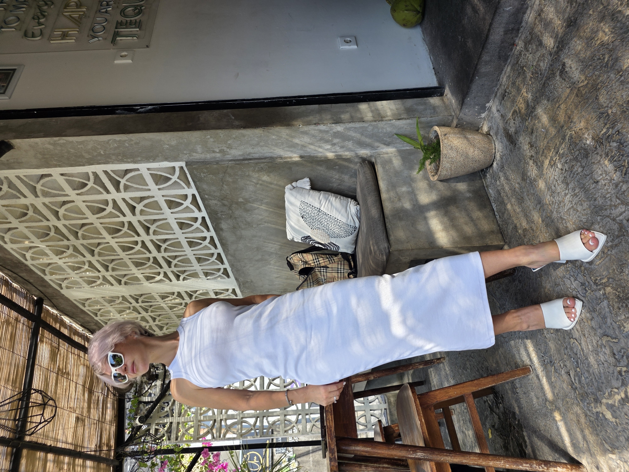 In North Kuta, Indonesia, a woman with short, light pink hair and white-framed sunglasses stands in a white, sleeveless, ribbed midi dress. She is wearing white slip-on heeled sandals. Her left arm rests on a wooden table next to her, and she has a bracelet on her left wrist and a ring on her left ring finger. Behind her, a padded bench is covered with a beige plaid bag and a white decorative pillow with a black and white pattern. The background features a textured concrete wall with a decorative white lattice screen and a bamboo-covered ceiling. The ground is made of textured concrete. A small potted plant sits to the right of the bench. The scene appears to be an outdoor seating area of a cafe or restaurant.