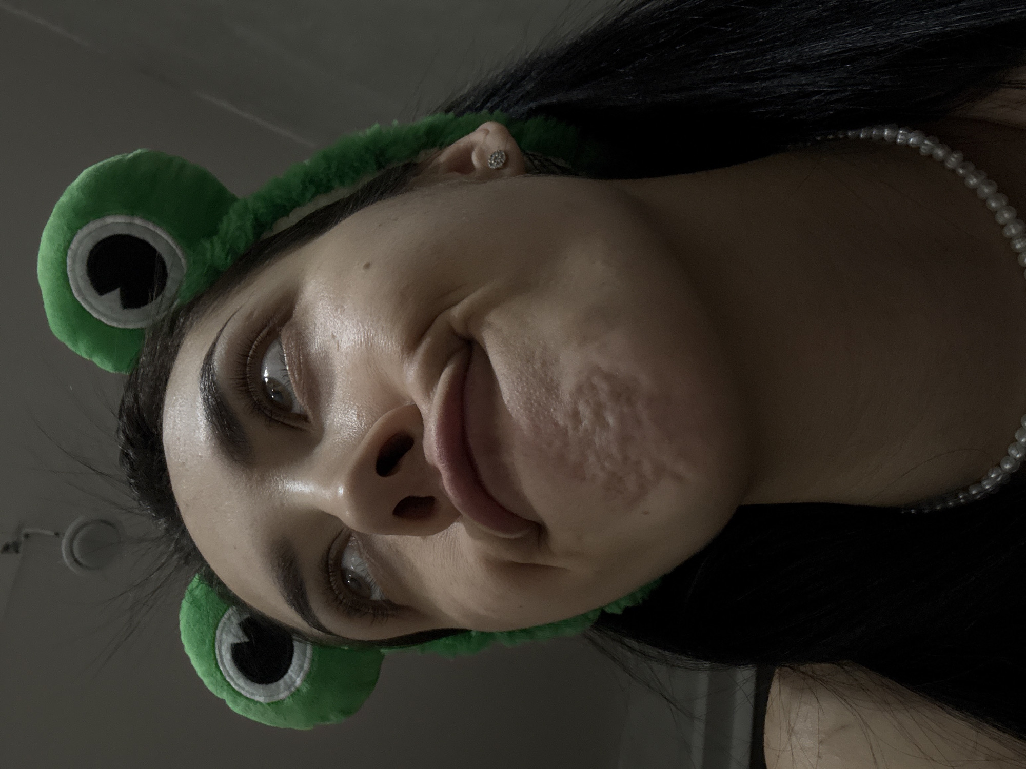 A close-up, low-angle photograph shows a person with dark hair and fair skin, wearing a green frog headband. The person is also wearing a pearl necklace and a stud earring in their left ear. The person's face is tilted downwards towards the camera, partially obscuring their neck. The background is an out-of-focus gray. There is no evidence to suggest that the image was taken in Votkinsk, Russia.