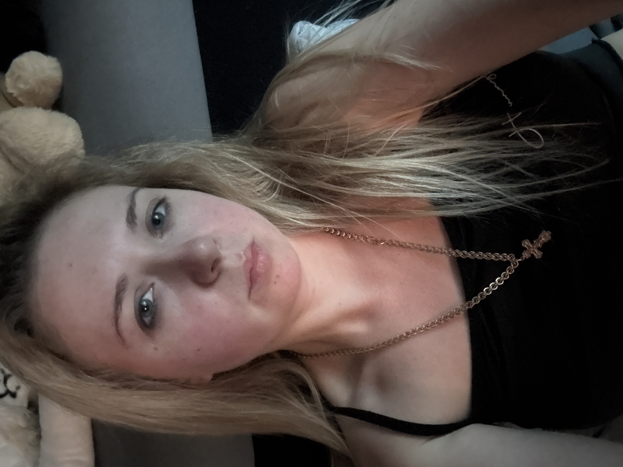 A woman with blonde hair, wearing a black tank top and a gold cross necklace, is looking at the camera. Her face is slightly flushed. Her hair is long and wavy. She appears to be indoors, with a dark background behind her. There is a stuffed animal in the upper right corner of the image.
