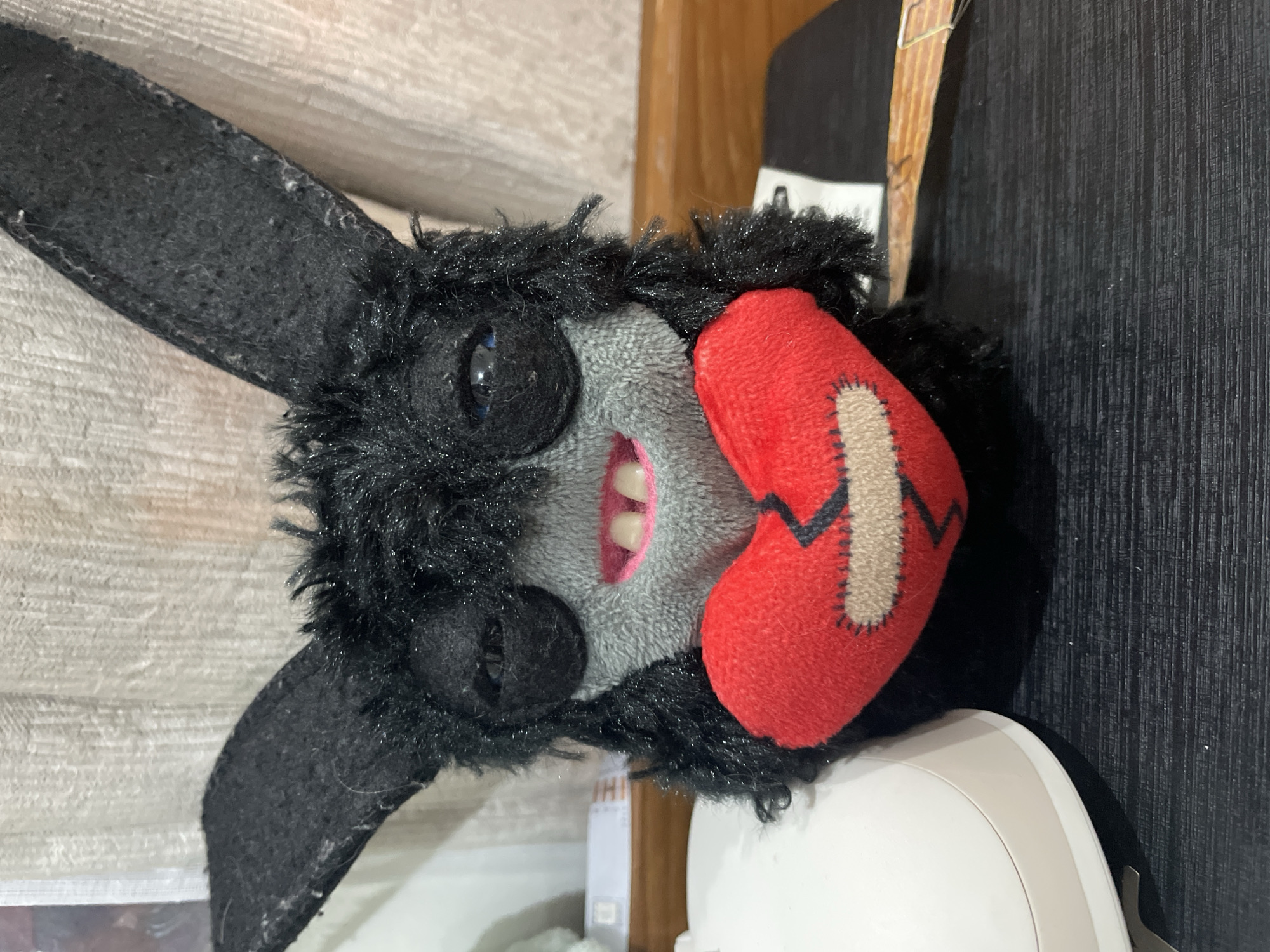 A plush toy rabbit with black fur and a grey face lies on its side. It has two black ears and visible white teeth. Attached to its head is a red heart-shaped patch sewn with a beige band-aid-like design. The setting appears to be indoors, with a wood-paneled wall and other objects in the background. There is no evidence to suggest a location within the city of Denpasar, Indonesia.