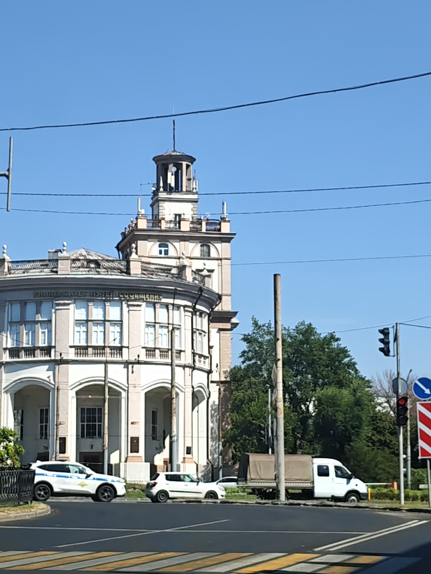 The image captures a street scene in daylight with a large, ornate building featuring a prominent tower. The building has a curved facade with numerous windows and arched entrances. A police car, a white van with a covered bed, and a smaller white car are visible on the street. Traffic lights and road signs are also present. The sky is clear and blue, and lush green trees are seen in the background. The overall impression is of an urban environment on a sunny day.