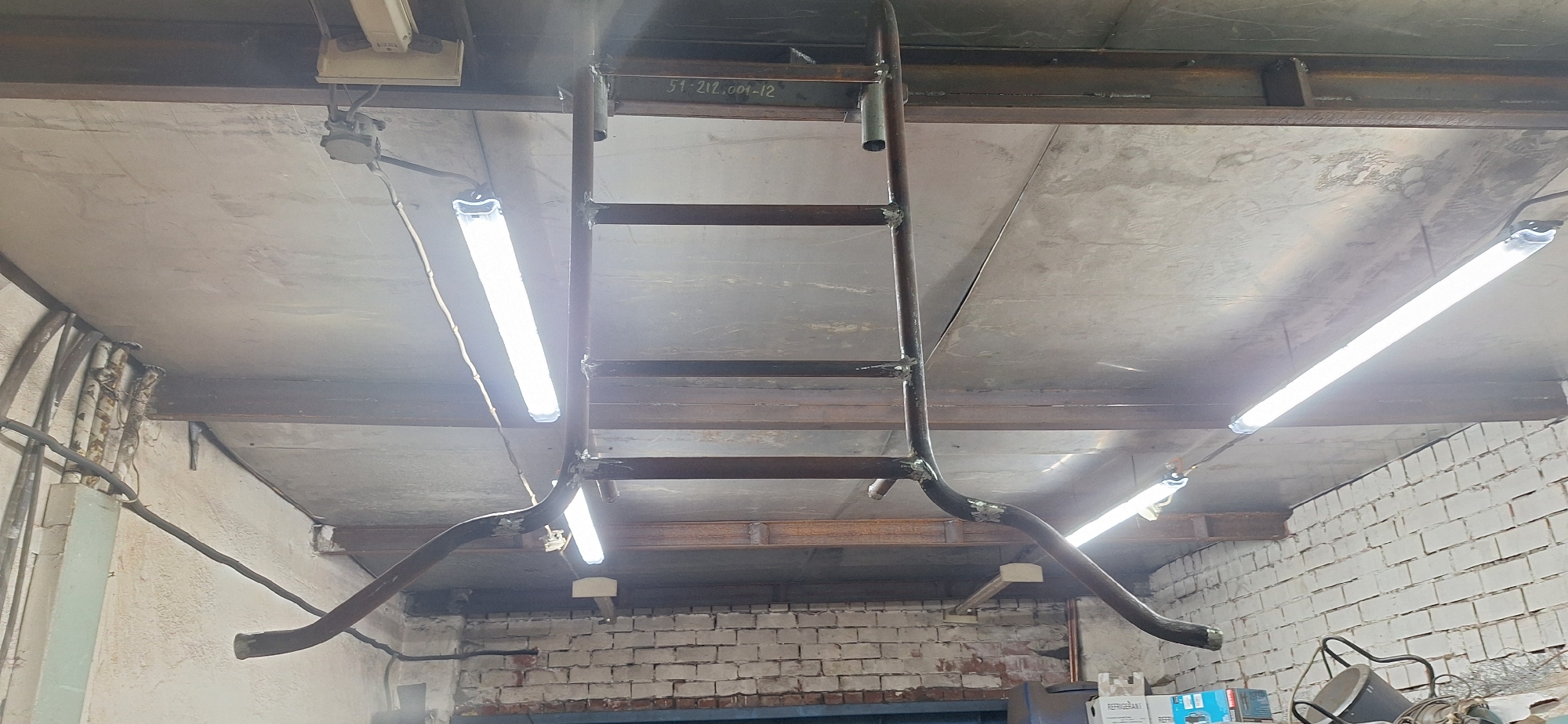 The image depicts an indoor scene, likely a workshop or industrial space, in Nakhodka, Russia. The focus is on a newly constructed metal structure resembling a ladder or scaffold, suspended from the ceiling. This structure appears to be made of welded metal pipes.
Two long fluorescent lights are mounted on the ceiling, illuminating the area. One light is on the left side of the frame, and another is on the right. The ceiling is a rough, grey concrete surface, supported by visible metal beams. The walls in the background are constructed of white brick. Electrical conduits and wires are visible running along the ceiling and walls, suggesting an active workspace.
The construction of the metal structure suggests an ongoing project or recent installation. The lighting is bright, indicating it could be daytime or the artificial lighting is sufficient for work. There is no visible human presence in the image.
A piece of text is faintly visible on one of the metal beams near the top of the frame: "51-212.001-12". This is likely a label or part number related to the construction or equipment in the space. The overall impression is one of a functional, utilitarian environment.
