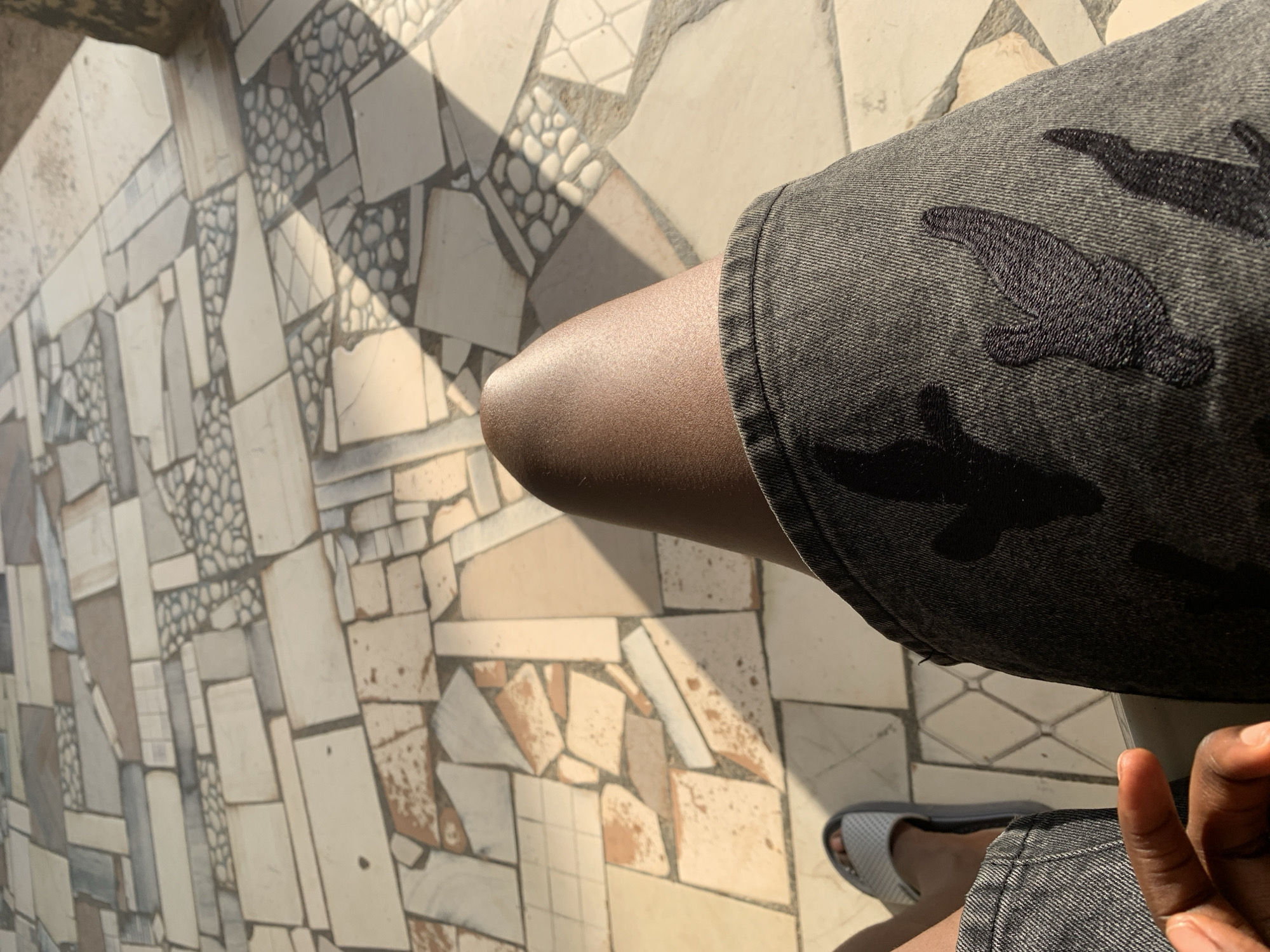 The image, captured in New Stadium, Nigeria, provides a first-person, downward-facing perspective of a person seated, focusing on their lap and the ground directly in front of them. The subject's right thigh, exhibiting dark skin, is centrally positioned and prominently illuminated by direct sunlight, creating a distinct highlight. The individual is wearing dark grey denim shorts or trousers, visibly adorned with multiple black embroidered flame-like or abstract patterns on the fabric of the right leg. On the far left side of the frame, the lower portion of the person's left leg is visible, with the left foot wearing a light grey slider-style sandal featuring a grey sole. The fingers of the left hand are briefly visible in the bottom left corner. The ground beneath is an elaborate mosaic or irregularly tiled surface, composed of various shapes and sizes of tiles in shades of off-white, beige, light brown, and some grey, interspersed with sections of small, light-colored pebbles or rounded tiles set in a darker matrix. Strong natural sunlight casts distinct, dark shadows across the tiled floor, including a prominent shadow of an unseen structure on the right side of the frame.