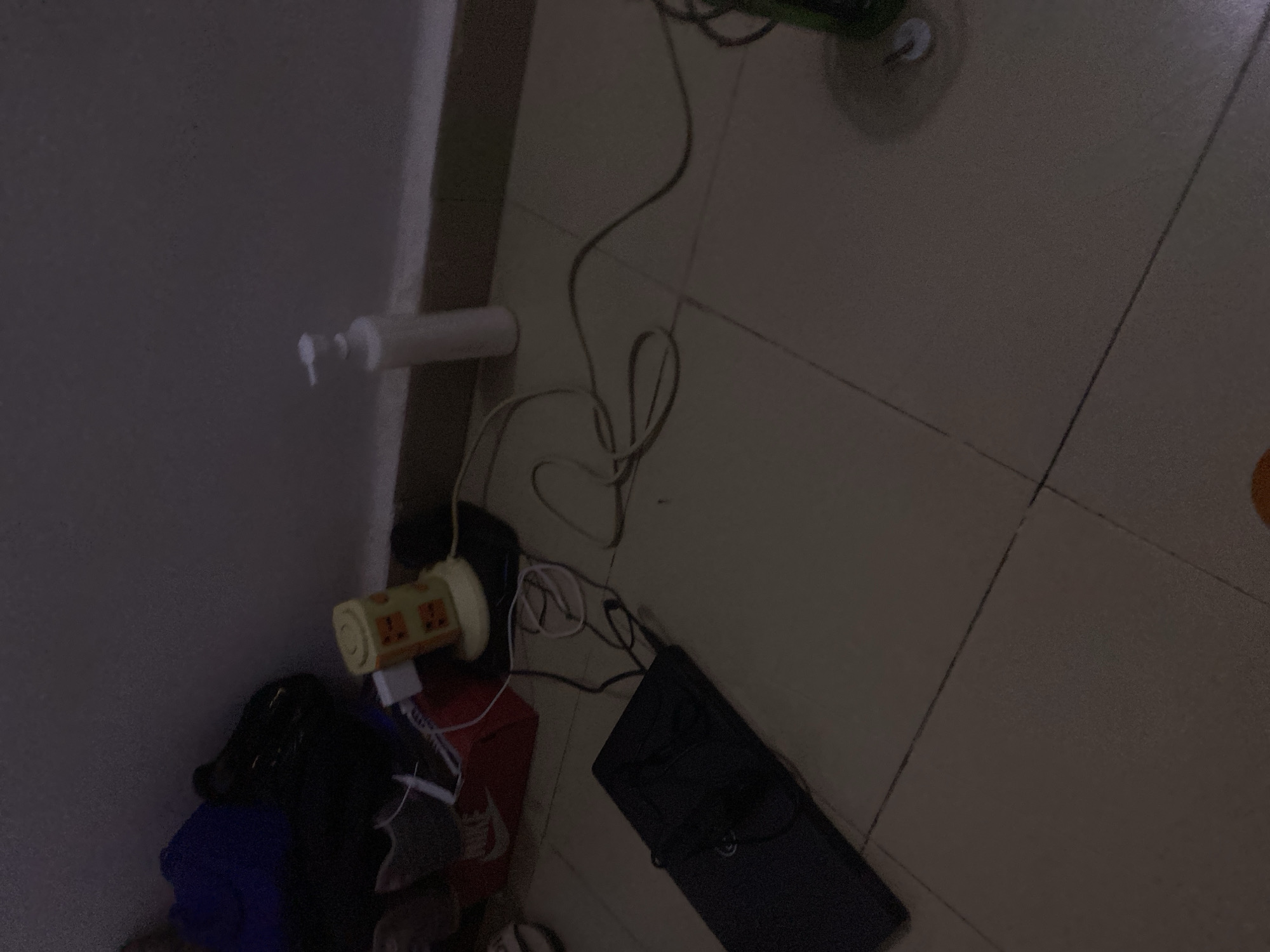 In Howayi, Nigeria, an image captures a dimly lit room with tiled flooring. The scene is cluttered with various objects, including a black laptop lying flat on the floor, connected to a tangled web of wires. To the left, a red Nike shoe box is visible, with a white shoe peeking out. Behind it, a black bag and a blue bag are partially obscured. Further to the right, a yellow object with orange accents, possibly a small fan or humidifier, sits on a black mat. A white dispenser bottle stands nearby. The room appears to be a personal space, likely a bedroom or study, given the presence of personal items and electronics. The overall ambiance is that of a lived-in space, with items scattered rather than neatly arranged.