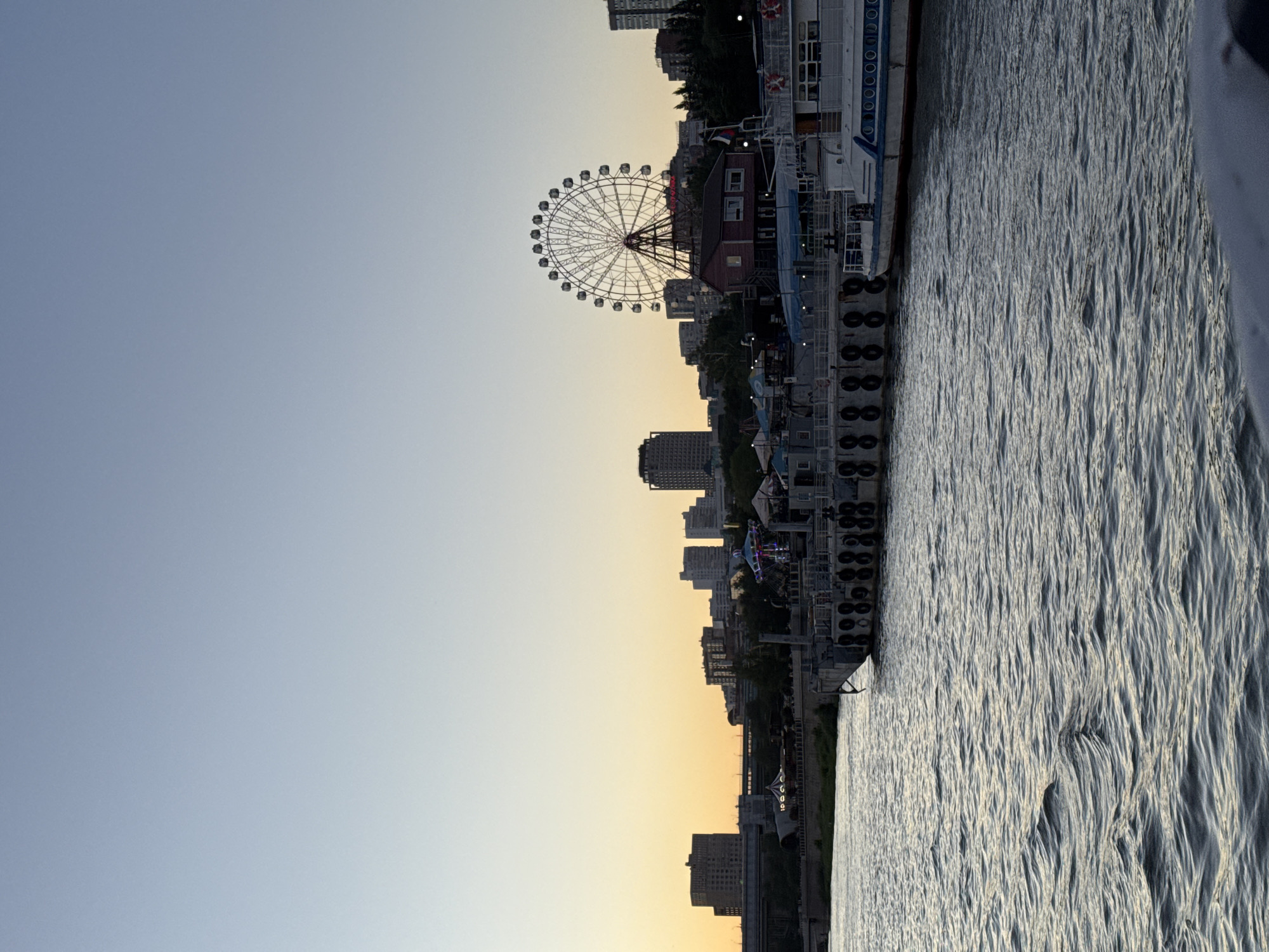 The image captures a waterfront scene in Novosibirsk, Russia, likely during dusk. A wide body of water with gentle ripples occupies the foreground, reflecting the pale light of the sky. In the mid-ground, a harbor area is visible, featuring several docked boats and a pier. One boat, partially visible on the right, appears to be a passenger vessel with white and blue detailing. The pier is lined with circular, dark-colored bumpers.
Beyond the harbor, the city skyline of Novosibirsk stretches across the horizon. Tall buildings of varying architectural styles dominate the background. Notably, a large Ferris wheel stands prominently, its spokes clearly defined against the sky, suggesting it is part of an amusement area. A small, red-roofed building is situated near the base of the Ferris wheel. The sky transitions from a pale yellow-orange near the horizon to a clear, light blue at the top, indicating the end of daylight. Trees and other vegetation are visible along the waterfront, interspersed with the urban structures. The overall atmosphere is serene, characteristic of an early evening by the water.