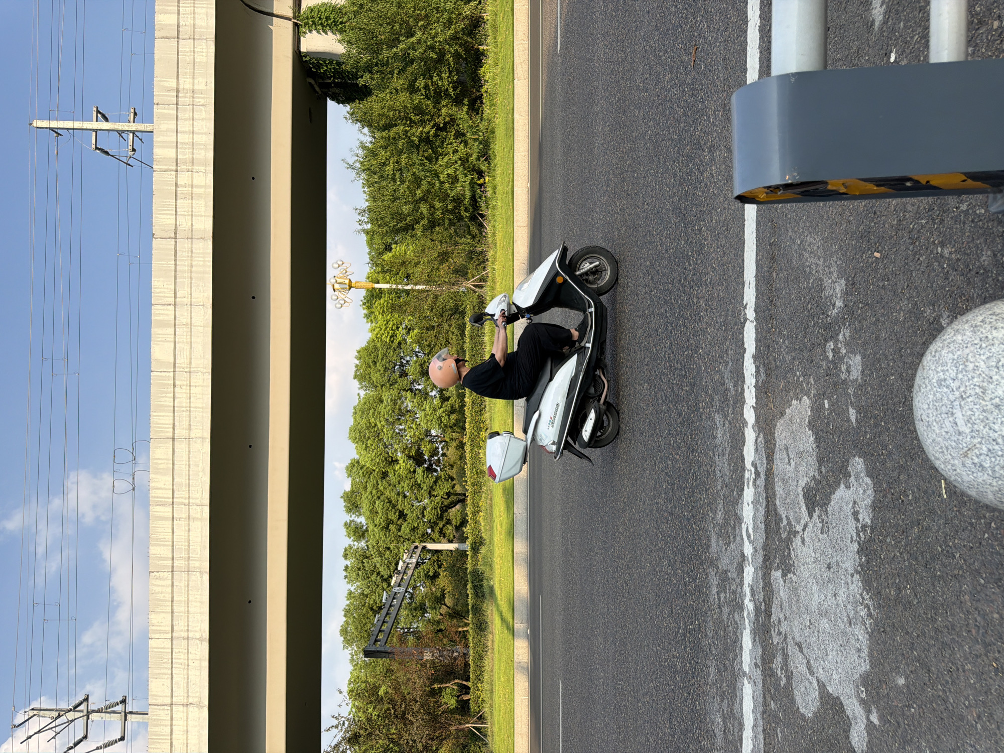 A person wearing a black shirt, black pants, and a light orange helmet rides a white and black scooter down a paved road in Jiangjin, China. The scooter has a white storage box on the back. The person appears to be looking forward. The road has white lane markings. In the background, there are green trees and a large concrete overpass with electrical wires running overhead. The sky is blue with some clouds. To the right of the road, there is a grey and yellow striped barrier.