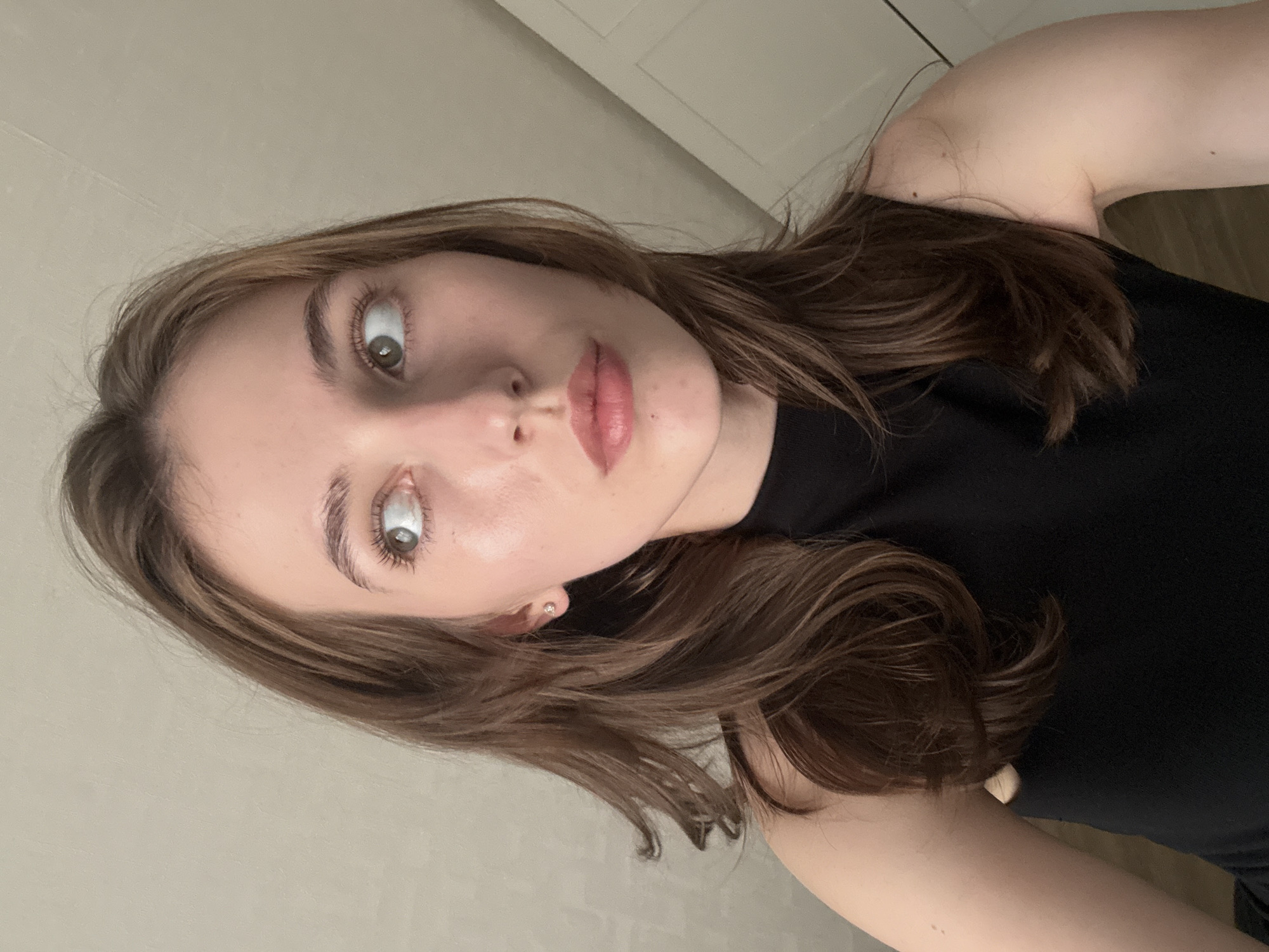 This is a close-up, medium shot photograph of a young woman looking to her left. She has light brown hair, styled in loose waves that frame her face. She is wearing a black, sleeveless top. Her skin appears fair and smooth. A small, stud earring is visible in her left ear. The background is a plain, light-colored wall, with what appears to be a door or panel on the right side. The lighting is soft and natural, suggesting an indoor setting. There are no discernible landmarks or features that would indicate a specific location within the city of Perm, Russia.
