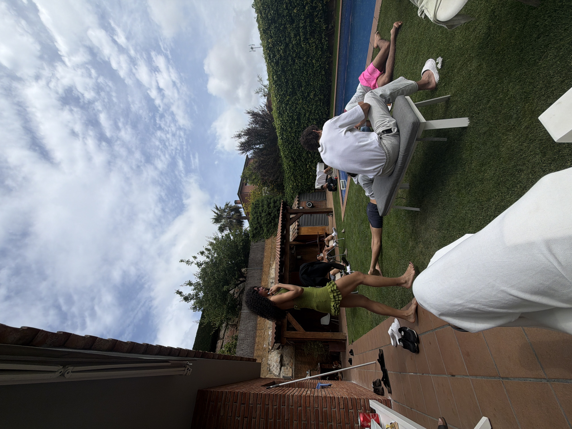 A group of people is gathered in an outdoor setting, likely a backyard in Sant Andreu de Llavaneres, Spain. There are individuals sitting on lawn chairs near a swimming pool, while others are standing and talking. A woman in a green dress is prominent in the foreground. The backyard includes grass, trees, a brick wall, and a building with a tiled roof. The sky is partly cloudy.