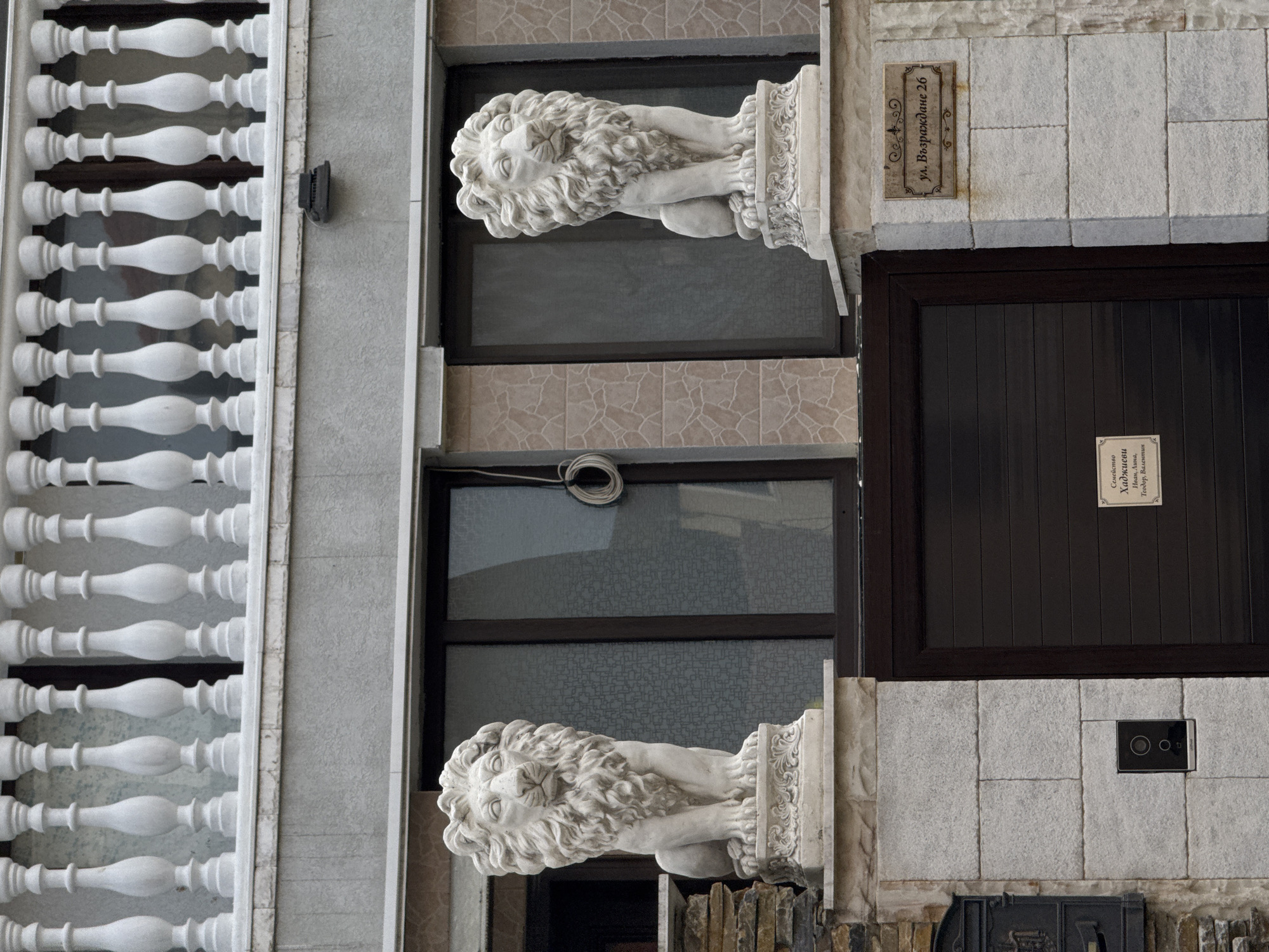 The image captures a portion of a building facade. Two lion statues, rendered in a classic style, flank a central window area. A balustrade of white balusters is visible on the left side of the frame. The address plaque indicates "ул. Възраждане 26," the address in the city of Burgas, Bulgaria. Below the right window, a rectangular plaque with Cyrillic script is affixed to the wall, reading, "Семейство Хаджиеви Иван, Лина, Теодор, Валентин" (Family Hadzhievi Ivan, Lina, Teodor, Valentin). A modern intercom system is also present near the bottom right. The building's exterior features a mix of stone and stucco textures.