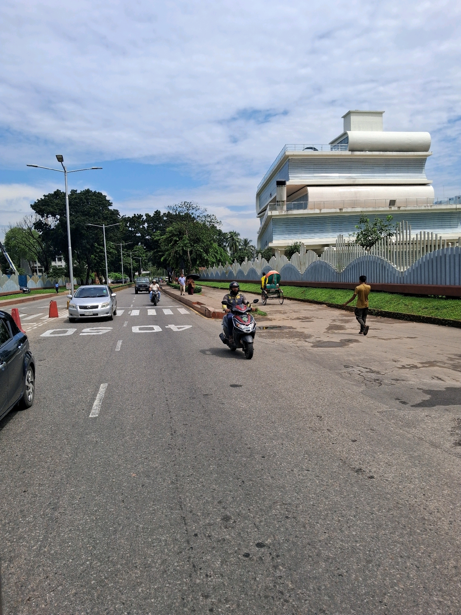 This image depicts a street scene in Dhaka, Bangladesh, on a cloudy day. The foreground features a paved road with white lane markings, including speed limit indicators "40" and "60". A silver sedan is stopped at a pedestrian crossing, with a red traffic cone nearby. To the right, a man in a yellow shirt and dark pants walks away from the camera, while further down the road, a person on a motorcycle approaches. A rickshaw is also visible further in the distance. The middle ground is characterized by lush green grass and a decorative white fence with wavy patterns. Beyond the fence, a modern, multi-story building with a unique, layered architectural design dominates the background. Tall trees line the left side of the road, and a lamppost stands tall on the left edge of the frame. The sky is partially covered with white clouds against a blue backdrop.